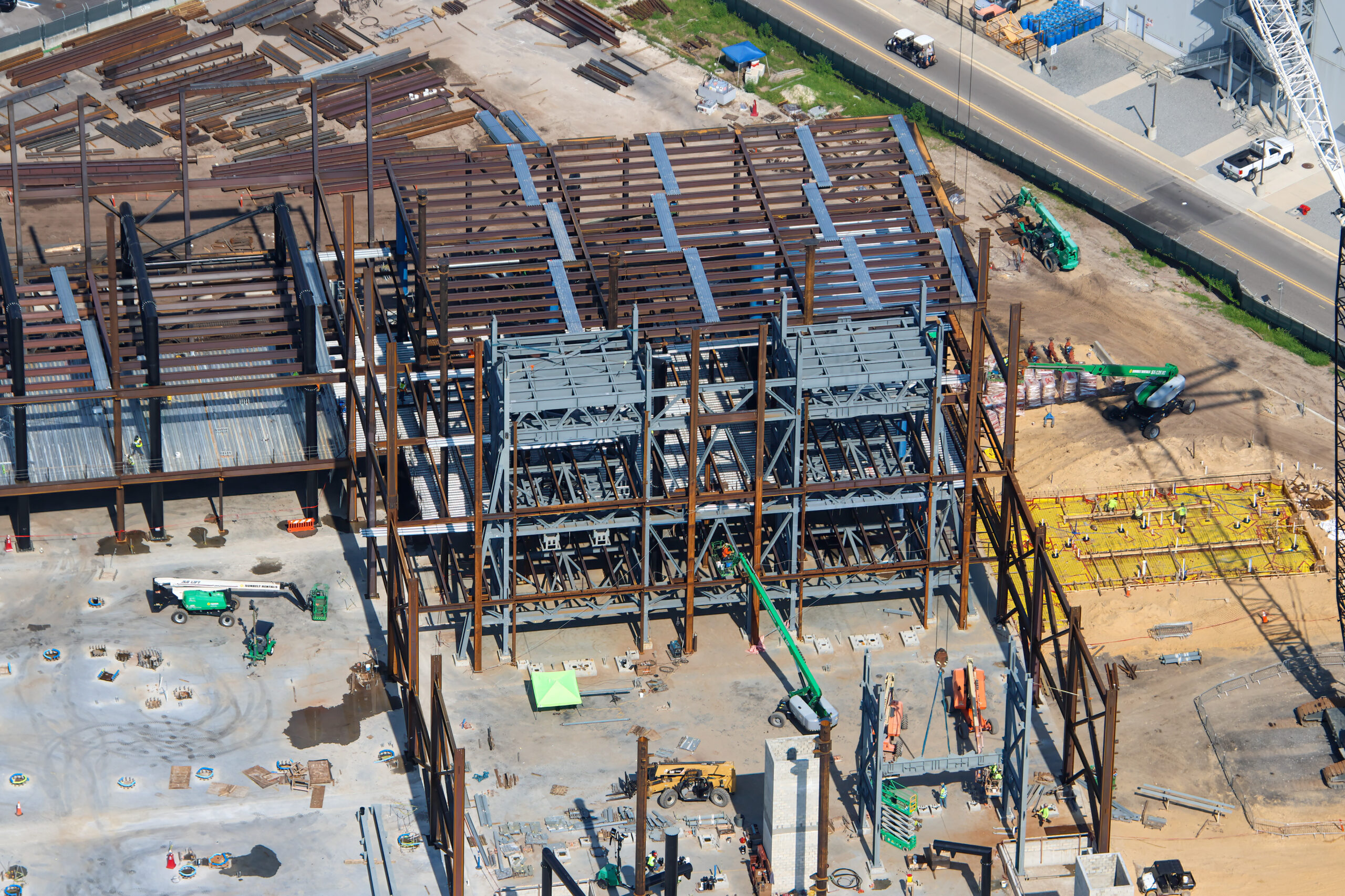 Overhead view of rust-colored structural steel columns inside the Monsters, Inc. coaster show building, with grey multilevel steel framing at center, corrugated floor decking laid across the bays behind it, and a yellow vapor barrier with rebar at right