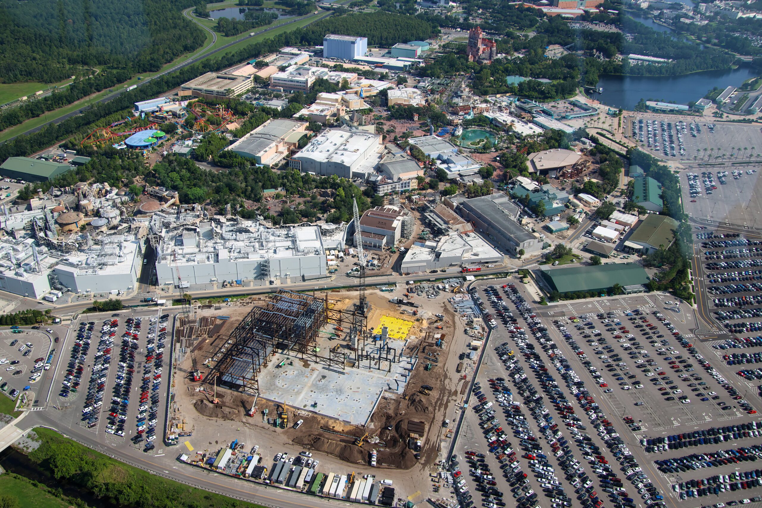 Wide aerial of Disney's Hollywood Studios showing the large Monstropolis construction site in the foreground, a dark steel frame rising from a pale concrete slab surrounded by dirt, cranes, and parking lots, with the pale show buildings of Star Wars: Galaxy's Edge beyond the road and its rockwork at left