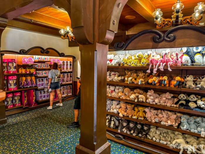 Interior of Der Teddyb&auml;r showing wooden shelves stacked with plush teddy bears, pandas, penguins, flamingos and other stuffed animals on the right, with a pink and purple Disney princess merchandise display on the left where a guest is browsing.