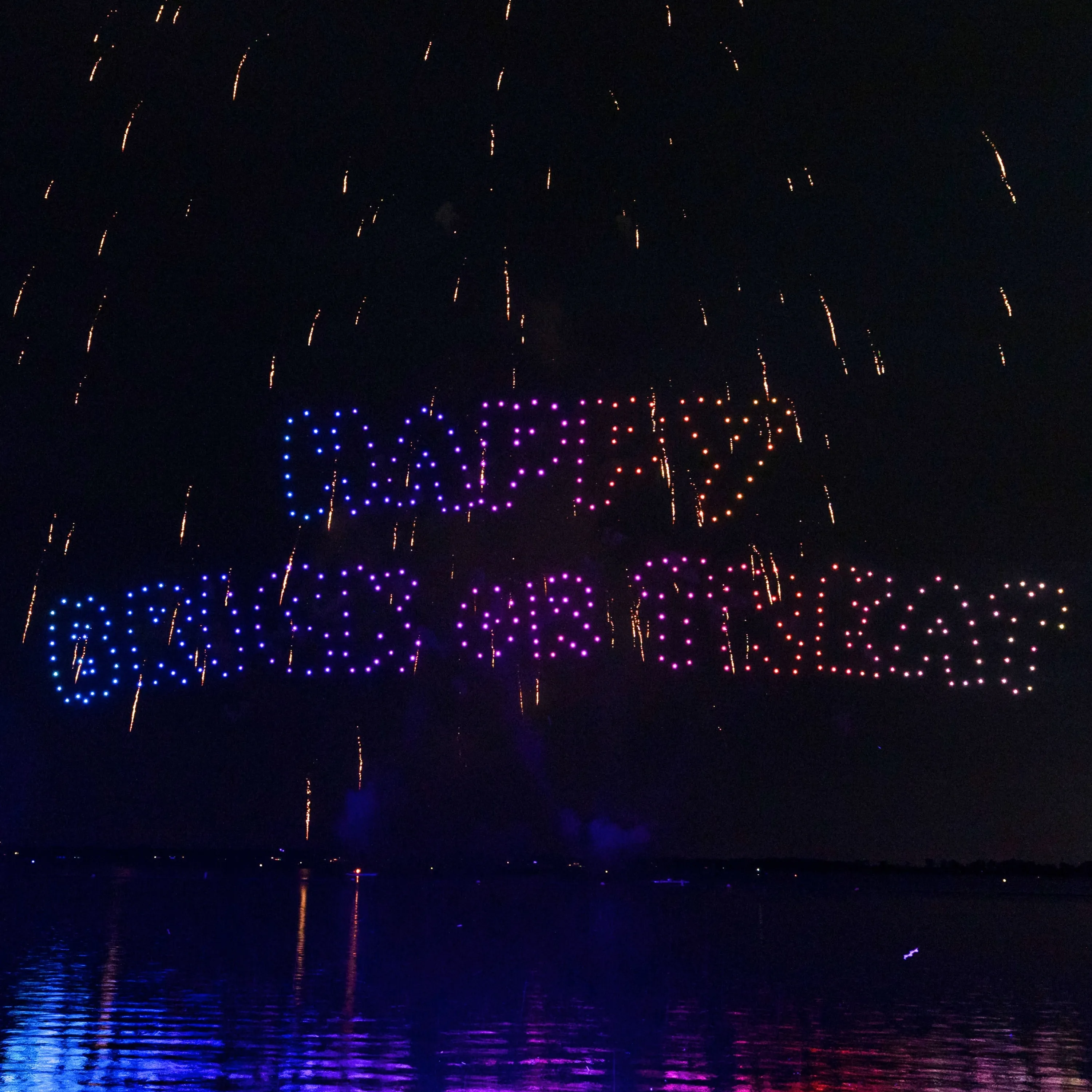 A drone show spelling out illuminated text in blue, purple and red lights above a lake, with firework streaks falling around the lettering and colored reflections on the water below.