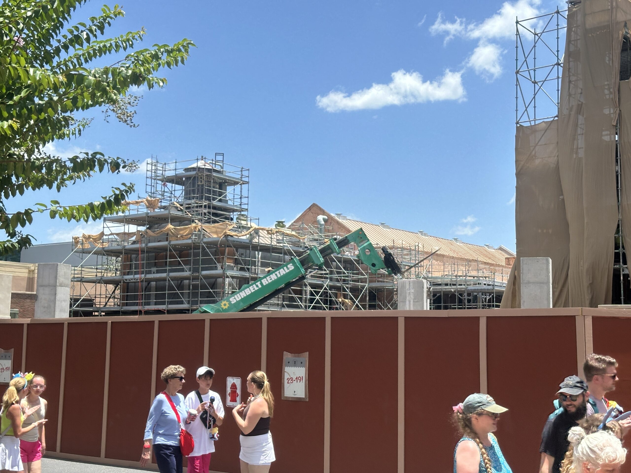 Ground level view from inside Disney's Hollywood Studios on a sunny day, with guests walking past a tall red construction wall. Behind it, pale concrete columns stand above the wall line, a green Sunbelt Rentals telehandler boom is raised, and a scaffolded show building with a wooden Brown Plumbing water tower rises in the background