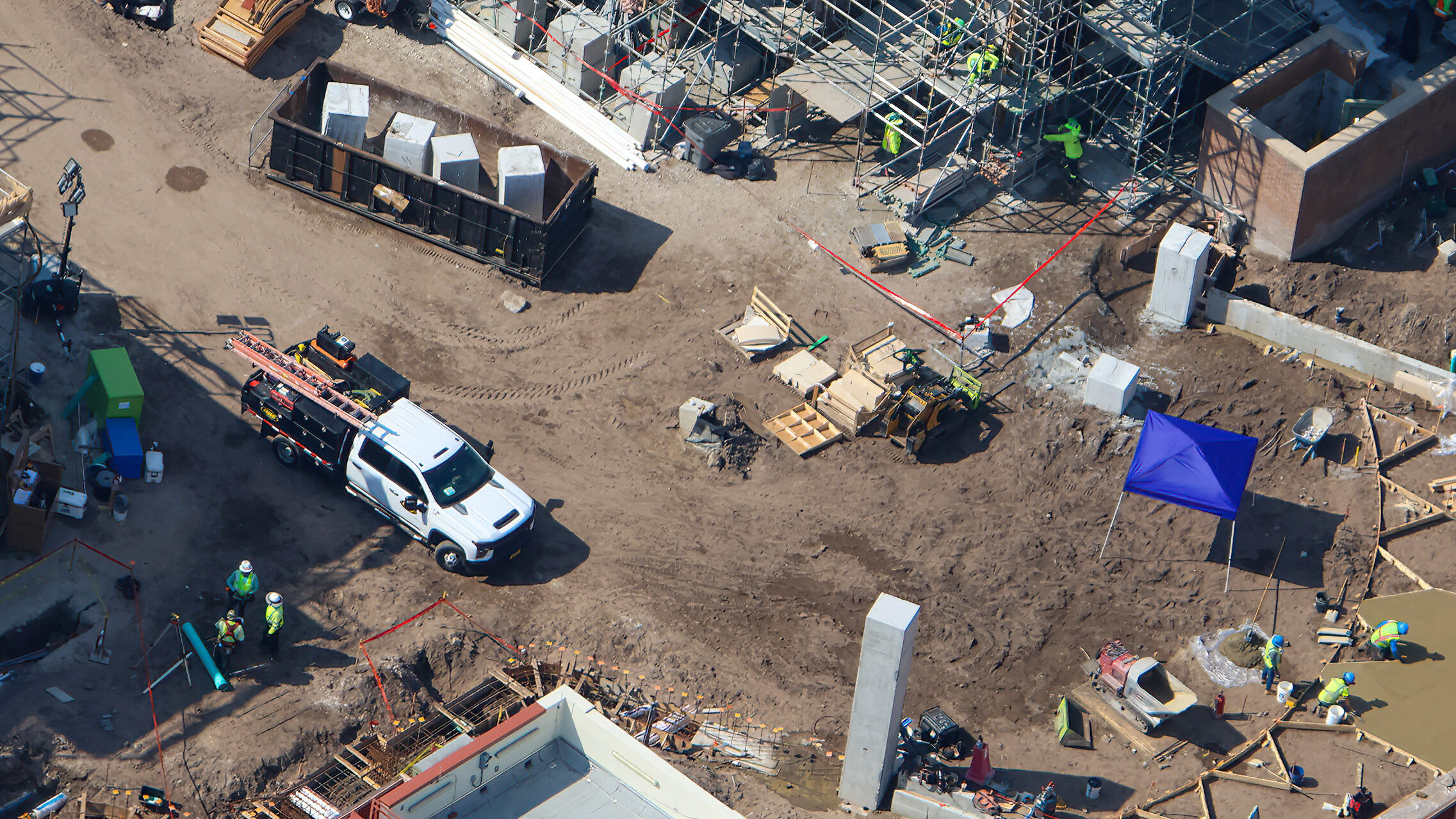 Close aerial view of the Monstropolis construction site showing a black roll-off dumpster holding four pale rectangular blocks of cut concrete, a white pickup truck with a ladder rack, hard-hatted workers in lime high-visibility vests, wooden concrete forms and a small yellow loader on bare dirt, a blue pop-up canopy, and one slim pale concrete column standing upright in the dirt at lower center