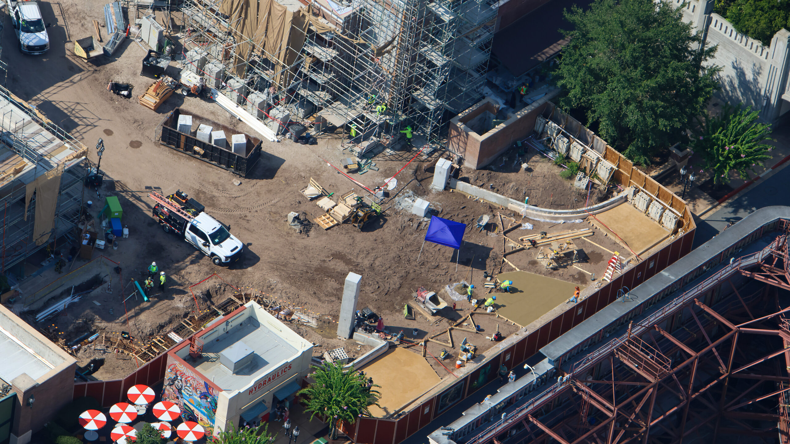 Wide aerial of the Monstropolis construction site showing large tan concrete slabs at right, with workers in lime vests and a concrete buggy around a smaller freshly poured section, curved wooden formwork and straight form boards laid out on bare dirt, a black dumpster and white pickup truck at left, rust red construction walls, and a building signed Hydraulics in Grand Avenue with red and white striped umbrellas in the lower left corner