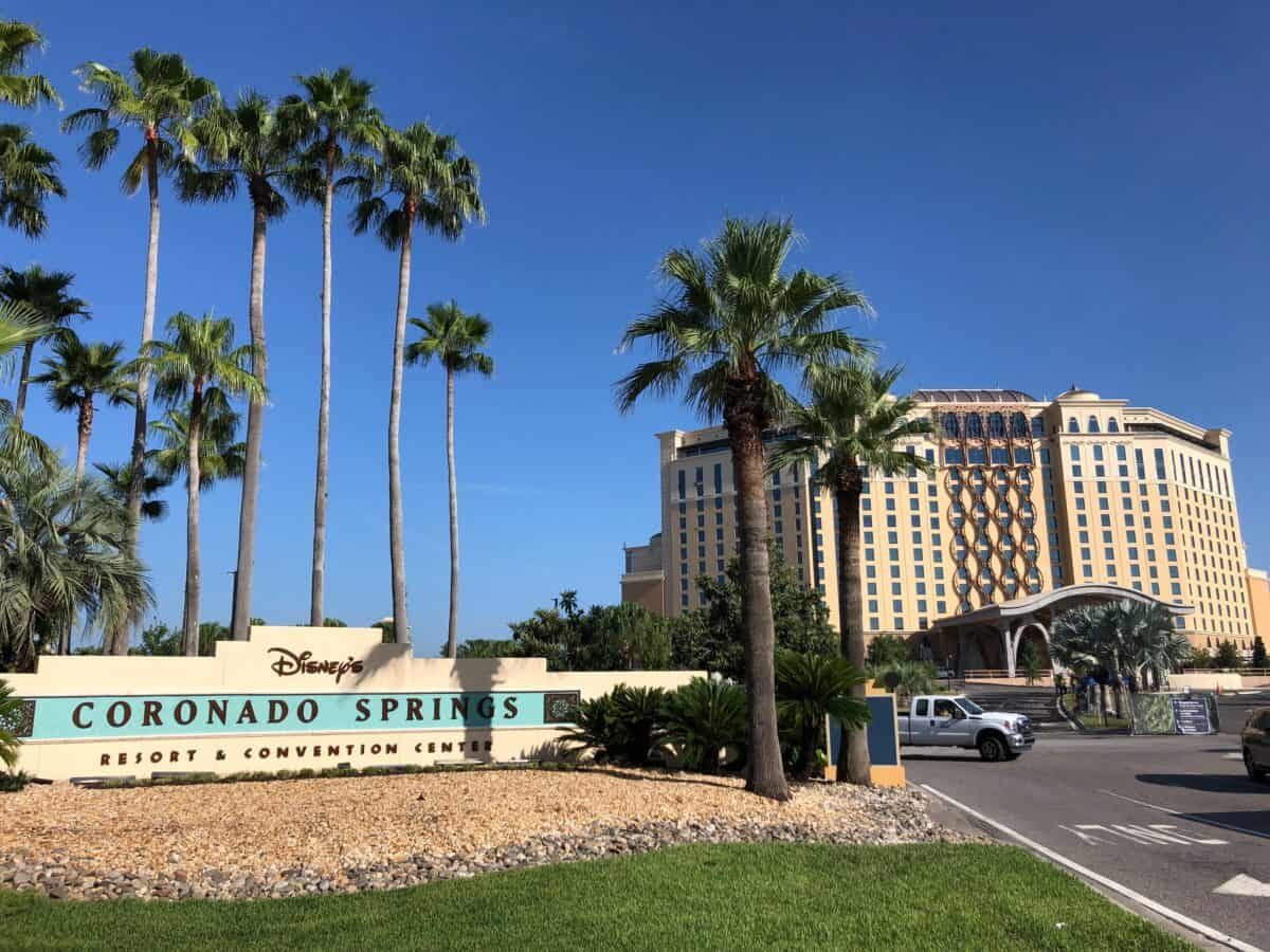 The cream stucco monument entrance sign with a teal tile band, reading Disney's Coronado Springs Resort and Convention Center, set in a bed of pale gravel with tall palm trees behind it, a green lawn in the foreground, and the cream and gold Gran Destino Tower rising against a clear blue sky in the background