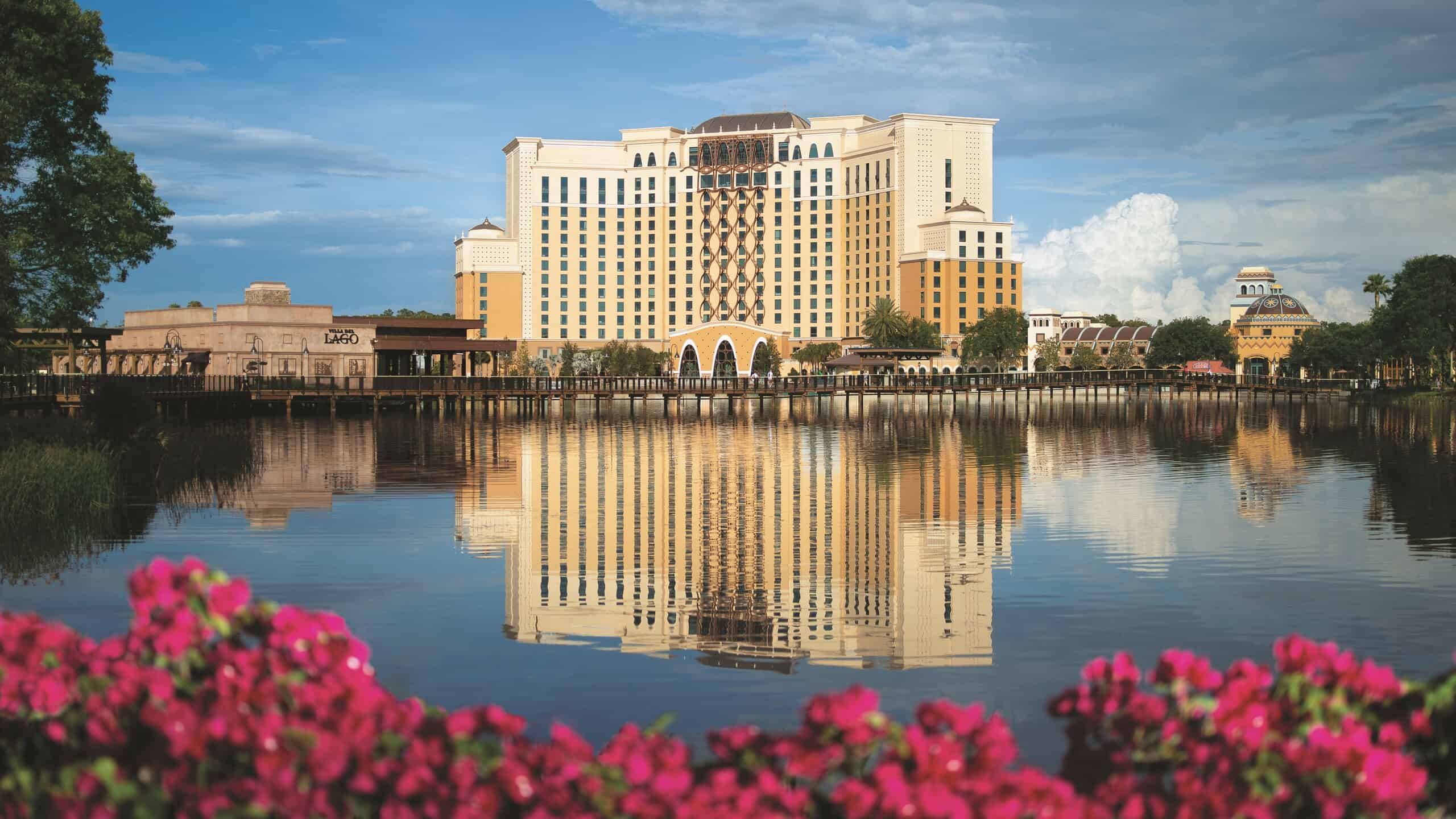 Wide daytime view across Lago Dorado at Disney's Coronado Springs Resort, with the tall cream and gold Gran Destino Tower rising in the center behind a long wooden boardwalk, the Villa del Lago building at left, palm trees and Spanish colonial rooflines along the shore, and bright pink bougainvillea blossoms filling the foreground