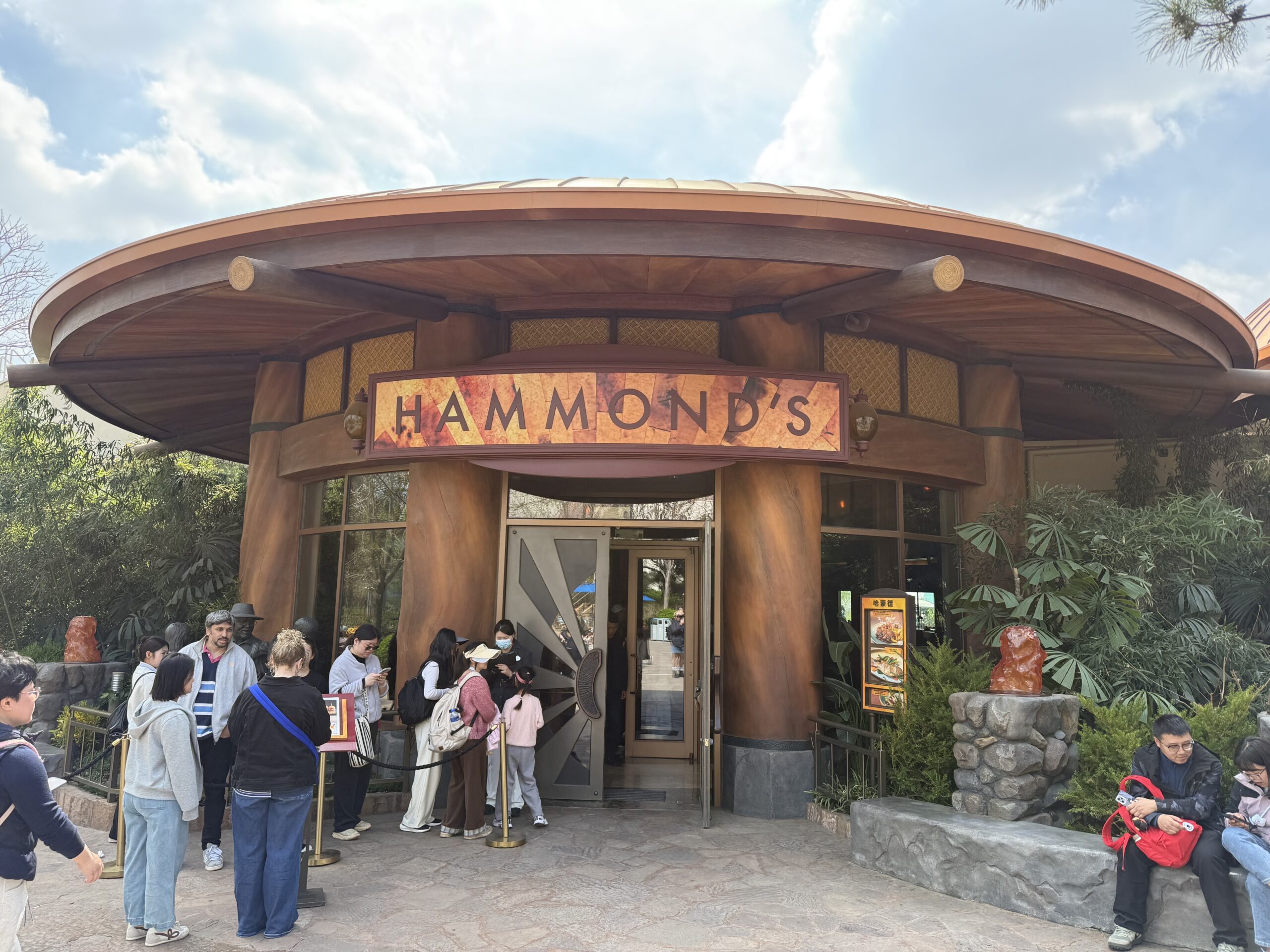 The entrance to Hammond's restaurant at Universal Beijing Resort, showing a wide circular roof supported by thick sculpted tree-trunk columns, an amber sign reading HAMMOND'S above the doorway flanked by two lantern sconces, a menu board with photos of dishes at right, and a line of guests waiting behind a rope stanchion at the door