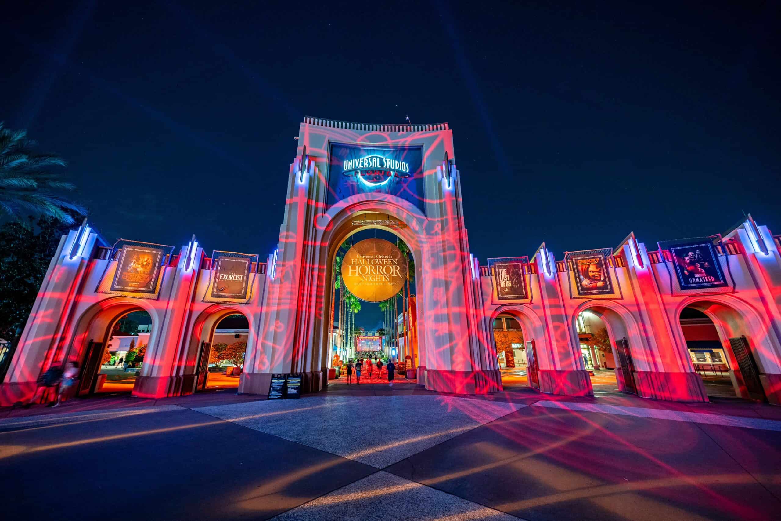 The Universal Studios Florida entrance arch at night, washed in red projected light with the illuminated Universal Studios Florida globe sign above and a round gold Halloween Horror Nights banner hanging in the central archway, flanked by rows of lit haunted house posters along the side arches, with guests walking through toward the park