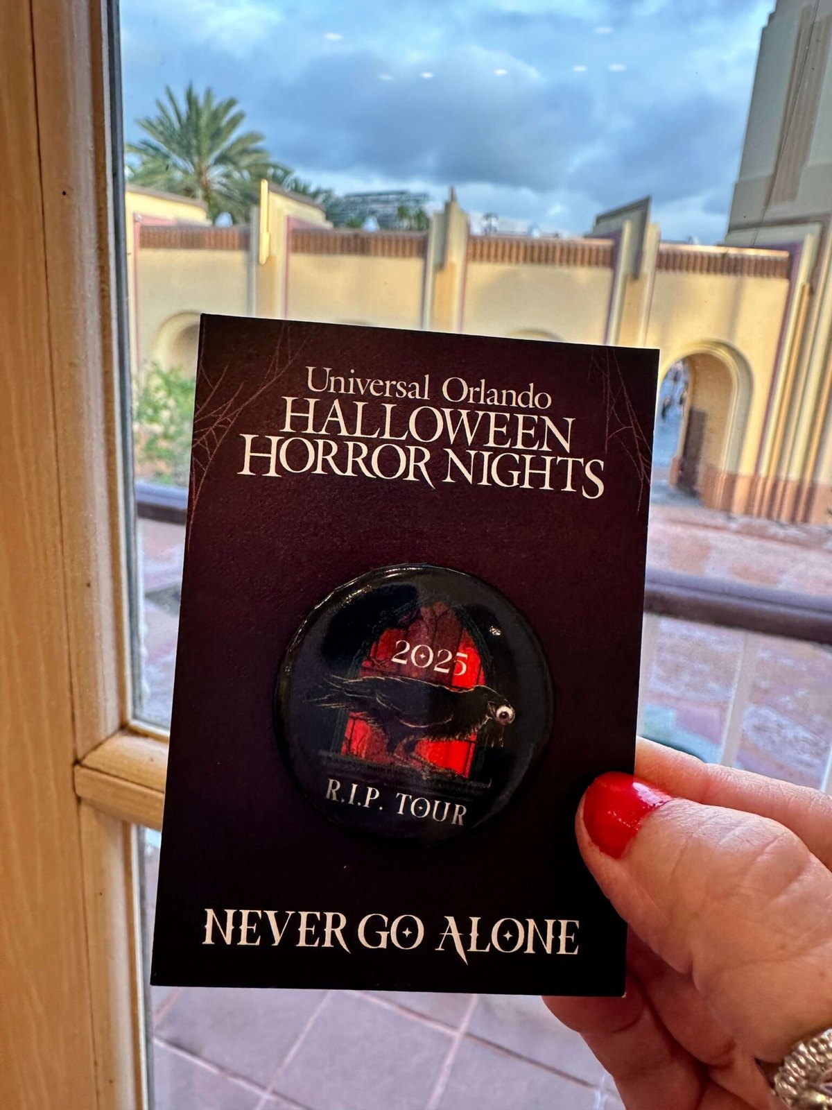 A hand holding a dark Halloween Horror Nights R.I.P. Tour card with a pinned round button showing a raven in a red-lit window, marked 2025, with the words Never Go Alone printed at the bottom.