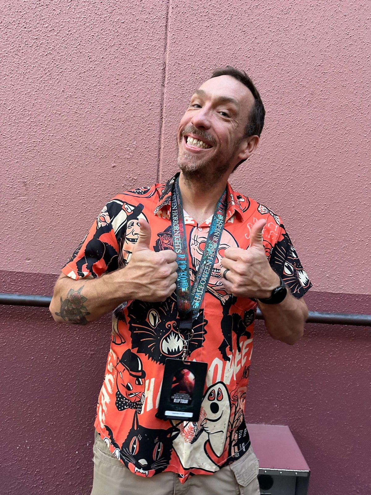 A smiling man in an orange Halloween-print shirt giving two thumbs up while wearing a dark Halloween Horror Nights R.I.P. Tour lanyard with teal lettering and a printed credential card hanging from it.