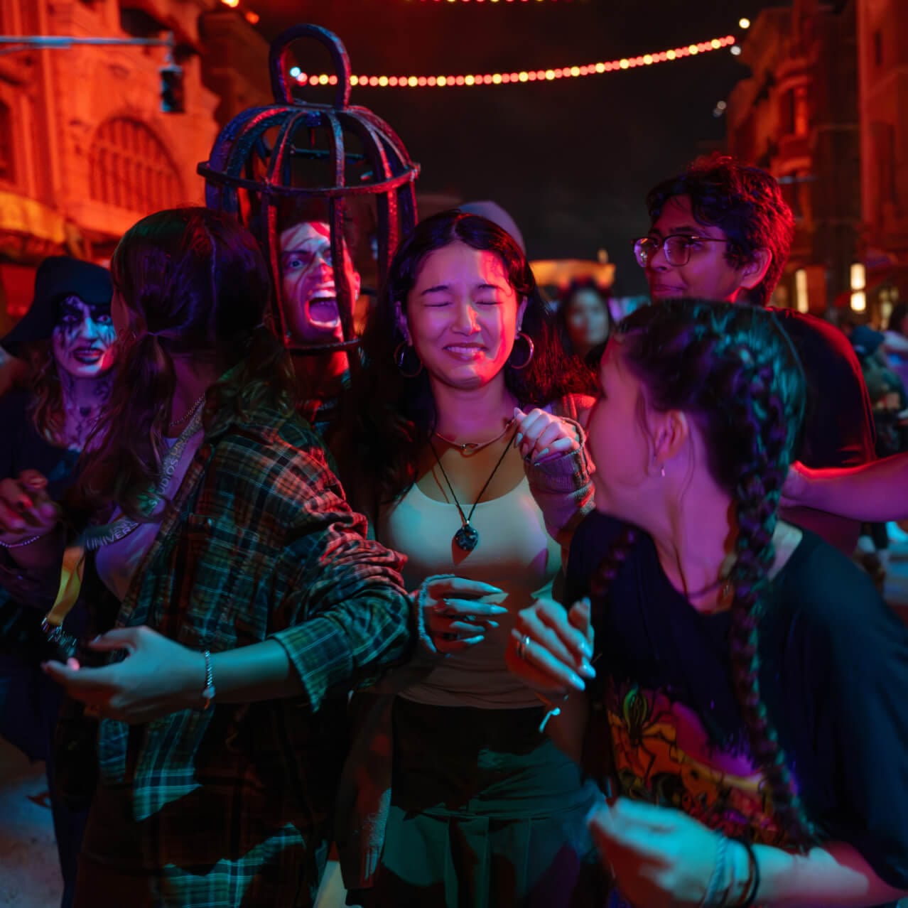 Guests in a Halloween Horror Nights street scare zone at night under strings of warm bulbs, laughing and recoiling as a scareactor wearing a metal birdcage over the head shouts at them, with another scareactor in white and black face makeup at the left edge and more guests crowding in around them, under red and teal lighting