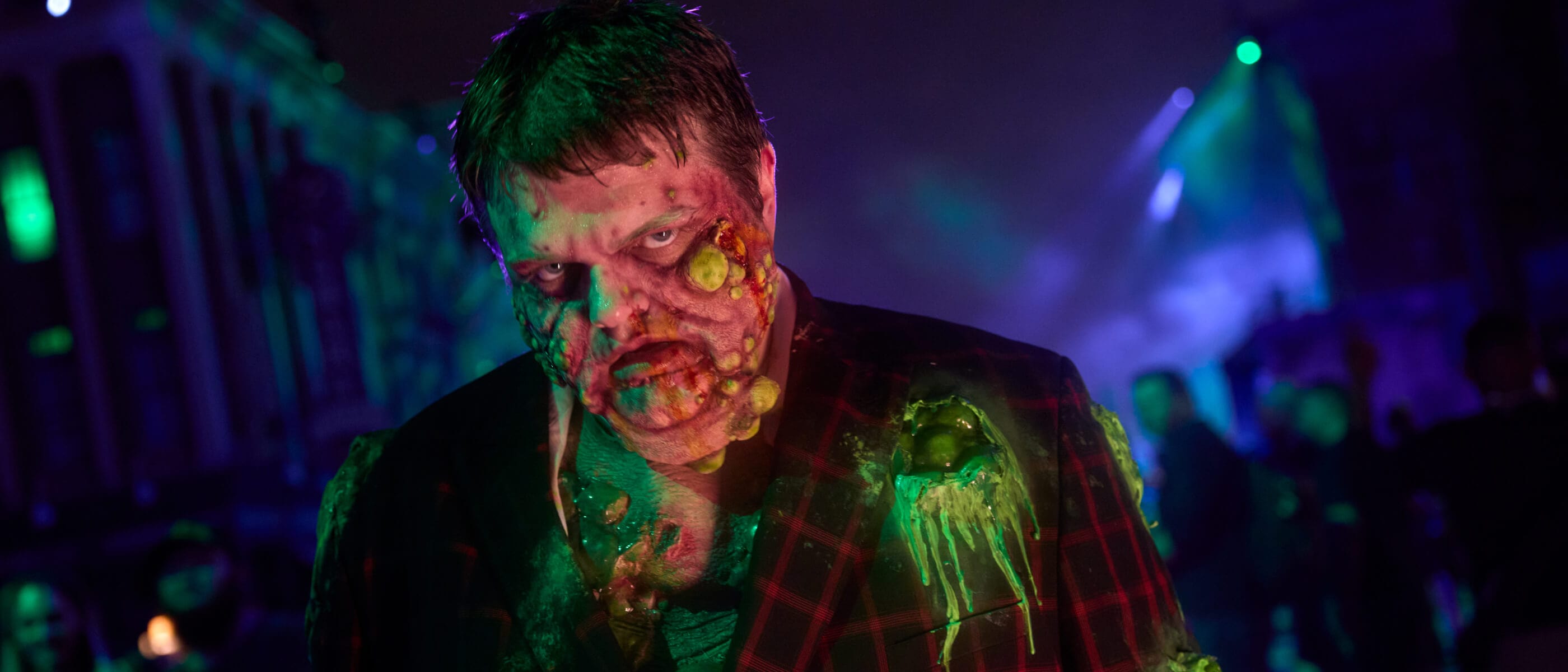 A Halloween Horror Nights scareactor lit in green and purple stage light at night, wearing a dark red plaid jacket, with heavy prosthetic makeup covering the face and chest in yellow-green boils and dripping slime, staring at the camera with blurred crowds and building facades behind