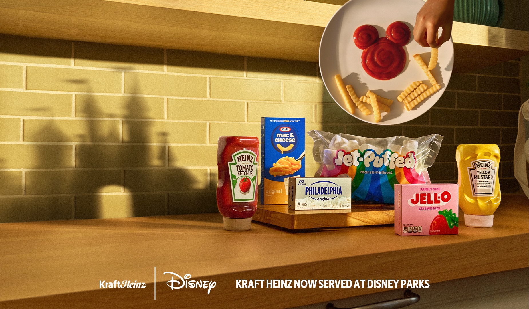 Campaign art showing HEINZ Tomato Ketchup, Kraft Mac and Cheese, Philadelphia cream cheese, Jet-Puffed marshmallows, strawberry JELL-O and HEINZ Yellow Mustard arranged on a wooden kitchen counter, with a castle shaped shadow falling across the tile wall behind them and a plate above holding crinkle fries and a Mickey shaped ketchup design, captioned Kraft Heinz Now Served at Disney Parks