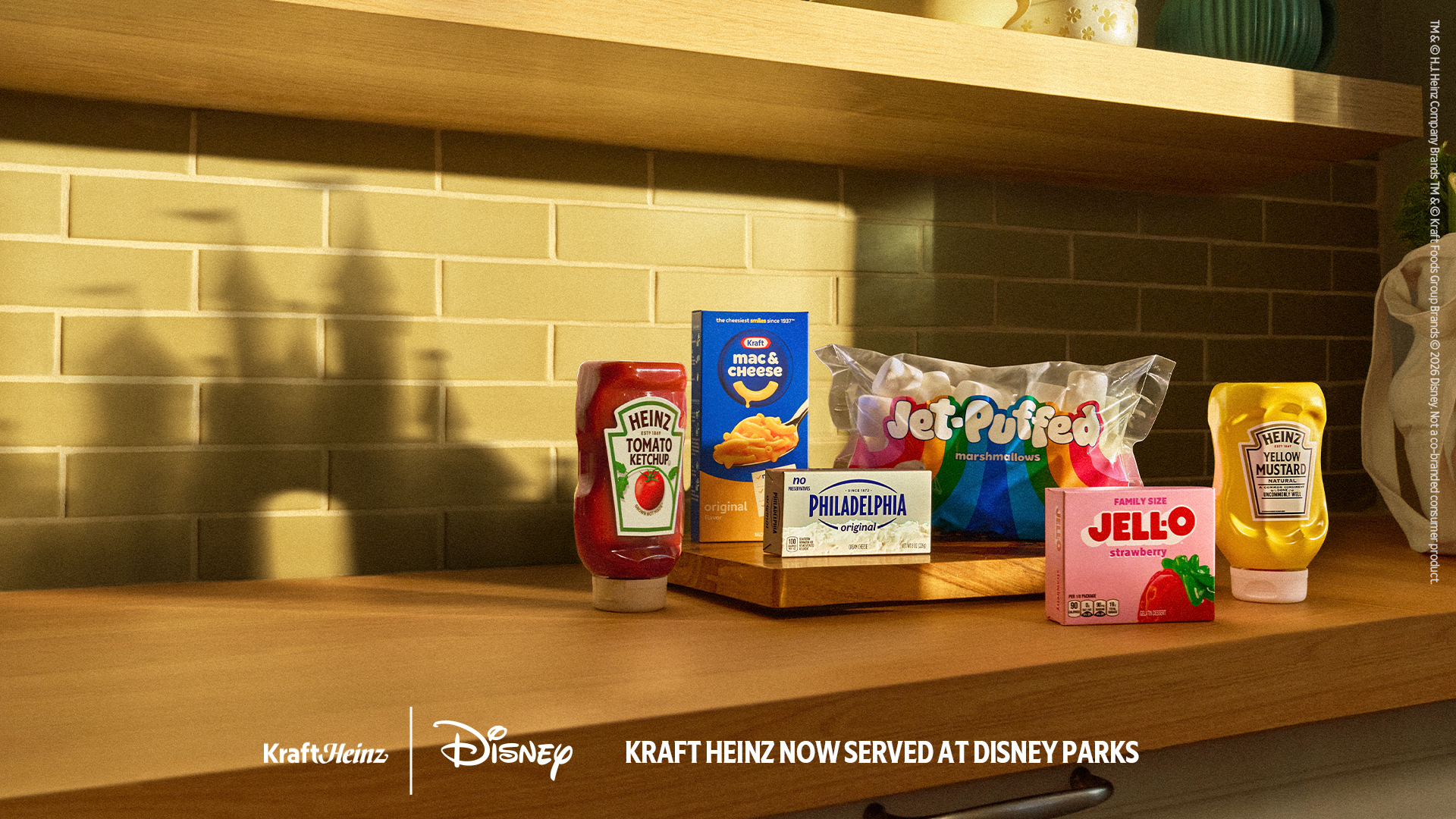 A second version of the same campaign art, showing the HEINZ Tomato Ketchup bottle, Kraft Mac and Cheese box, Philadelphia cream cheese, Jet-Puffed marshmallows, strawberry JELL-O and HEINZ Yellow Mustard on a wooden counter with a castle shaped shadow on the tile behind, above Kraft Heinz and Disney logos