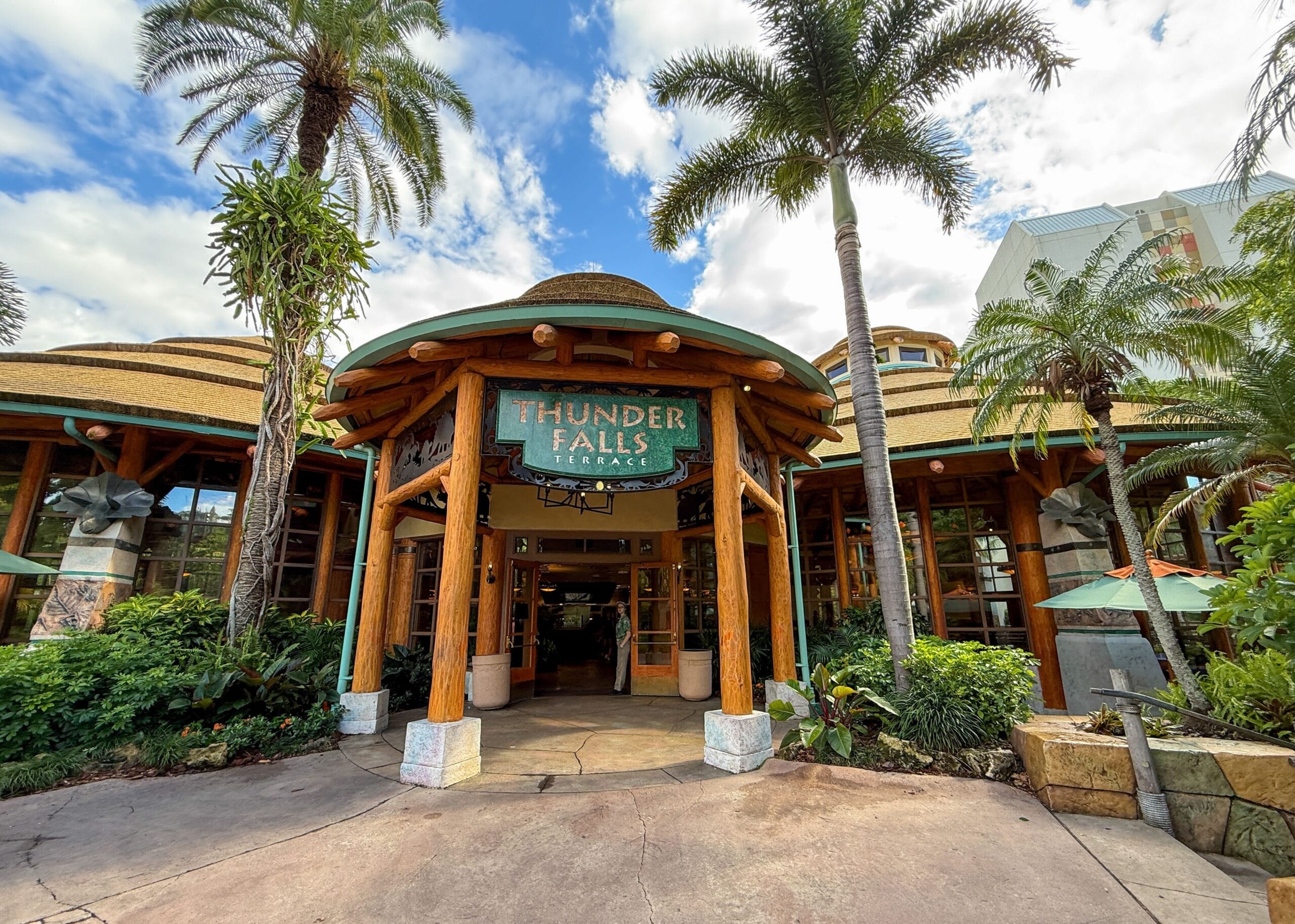 The entrance to Thunder Falls Terrace at Islands of Adventure on a partly cloudy day, showing a round thatched-roof pavilion supported by thick peeled log columns, a weathered green sign reading Thunder Falls Terrace hanging in the center, tall palm trees on either side, lush tropical planting, and a team member standing in the open doorway
