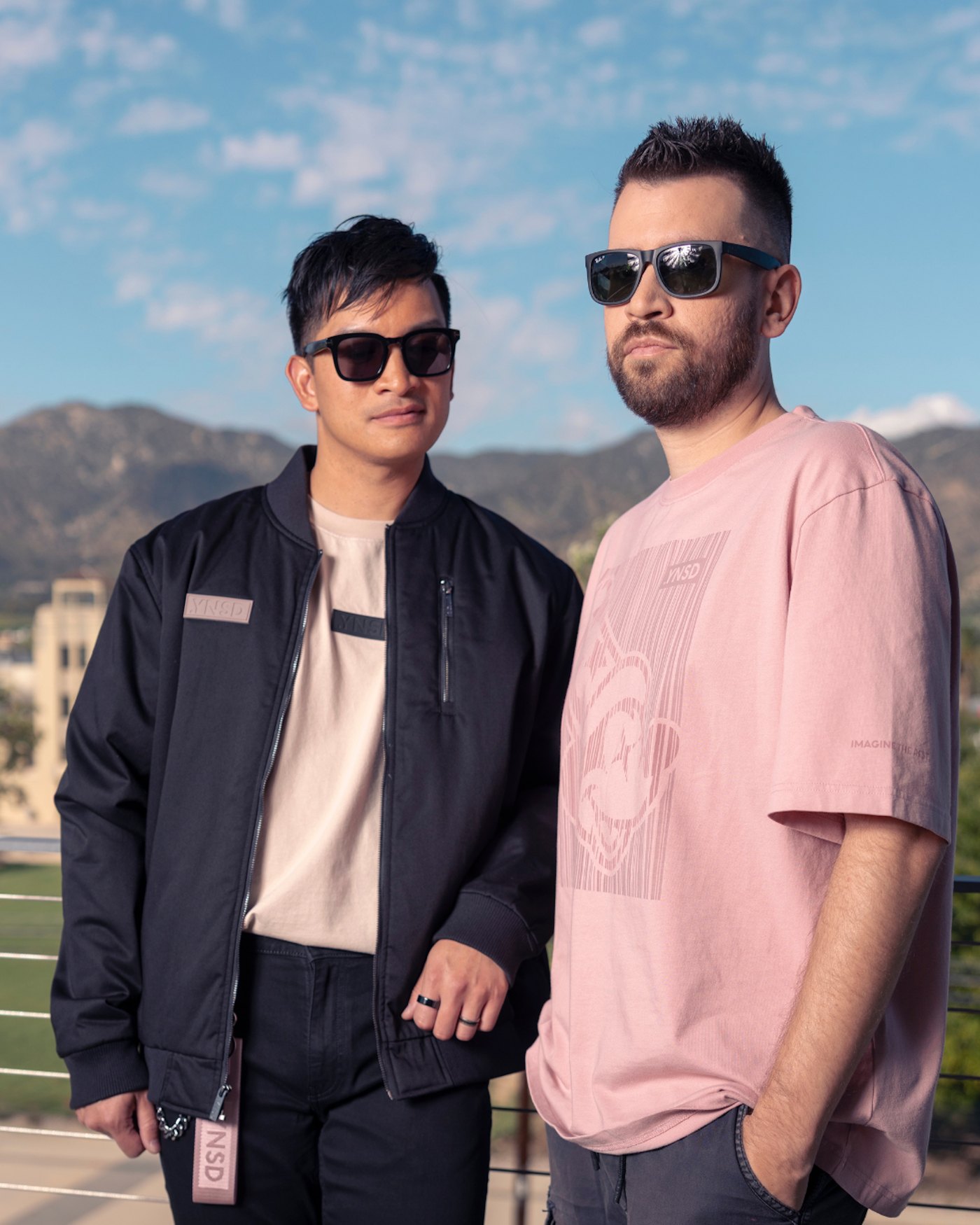 Two models on a rooftop, one wearing a dark bomber jacket with pale .YNSD patches and a long zip pull tag, the other in a dusty pink tee printed with a tonal graphic in the same shade as the fabric.