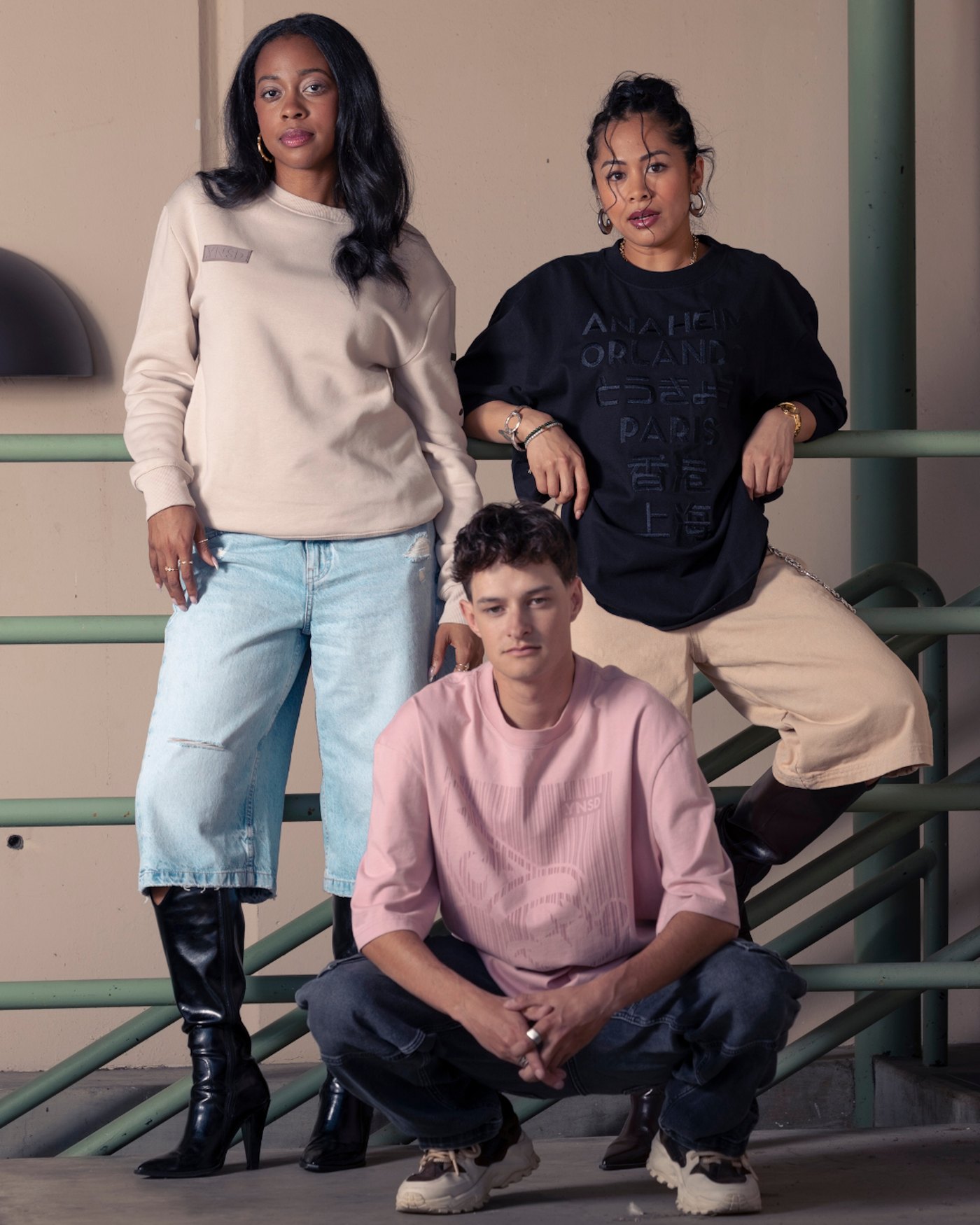Three models posed against a pale green metal railing wearing pieces from the collection: a cream crewneck with a small .YNSD box patch, the navy city-list tee, and a pink tee with a tonal printed graphic.