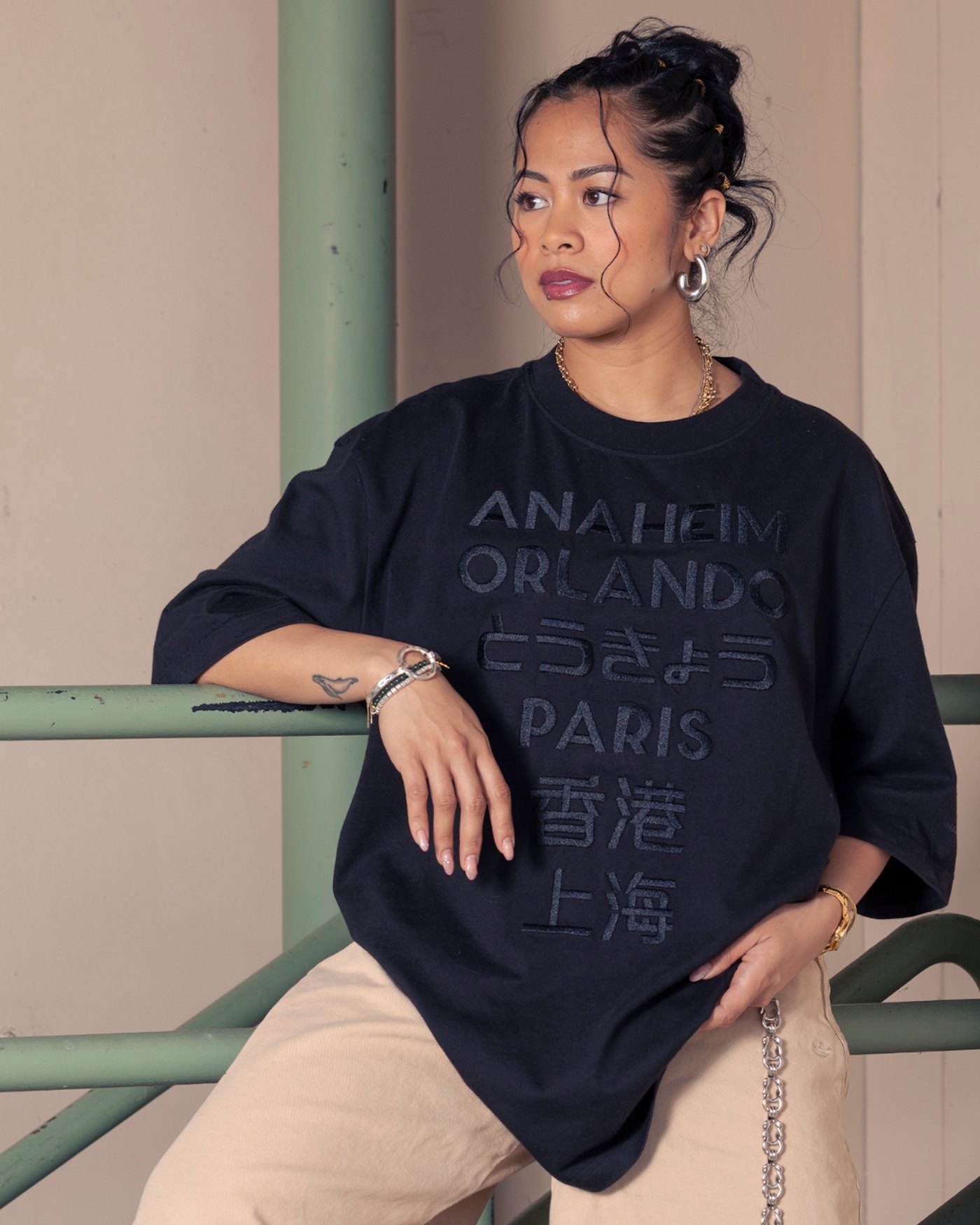 A model in an oversized navy tee embroidered in matching dark thread with a stacked list of city names reading Anaheim, Orlando, Tokyo in Japanese characters, Paris, and Hong Kong and Shanghai in Chinese characters.