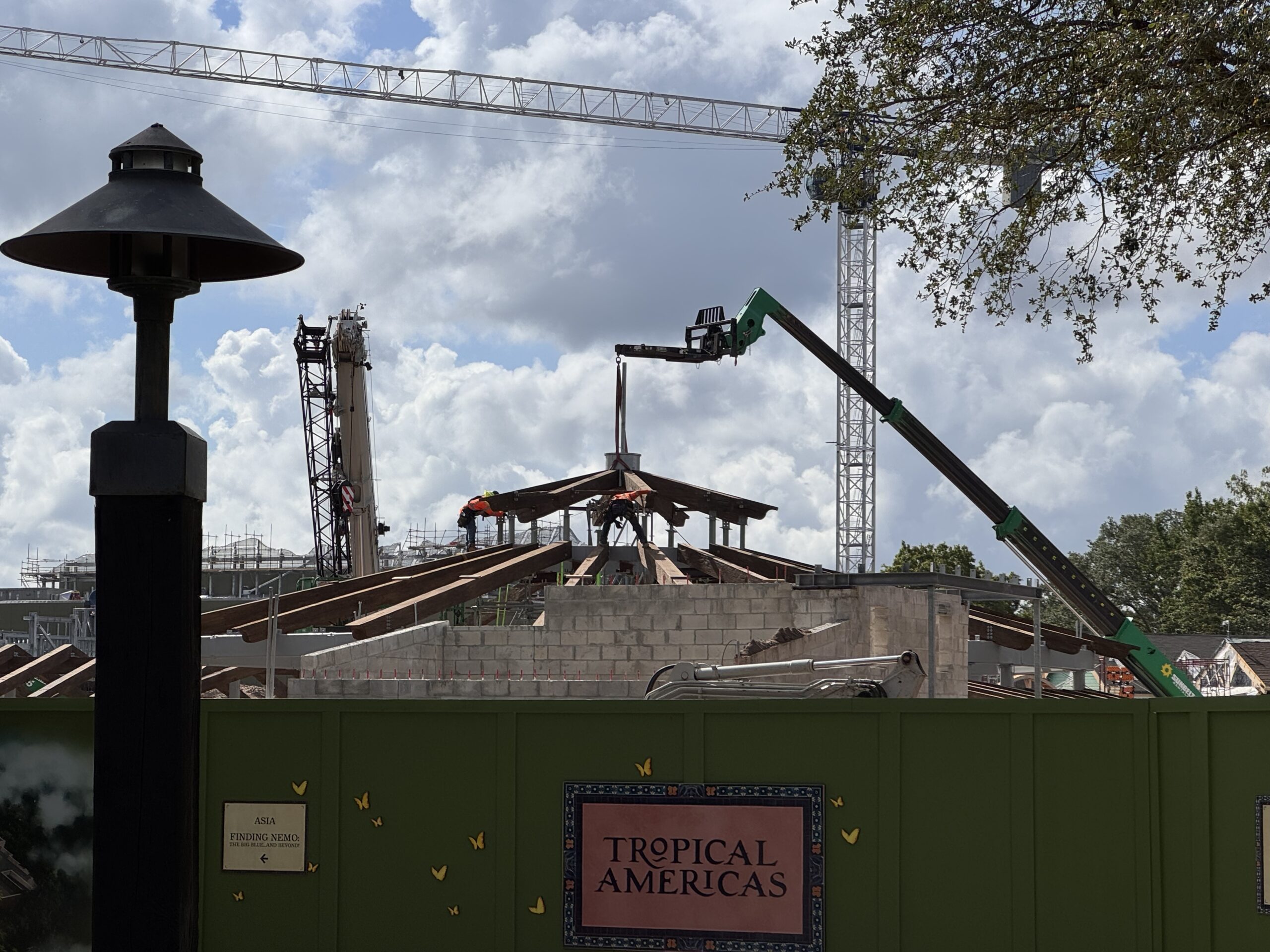A green boom lift lowers the roof framing onto the Tropical Americas carousel at Disney's Animal Kingdom as workers secure it, with a Tropical Americas sign on the construction wall below