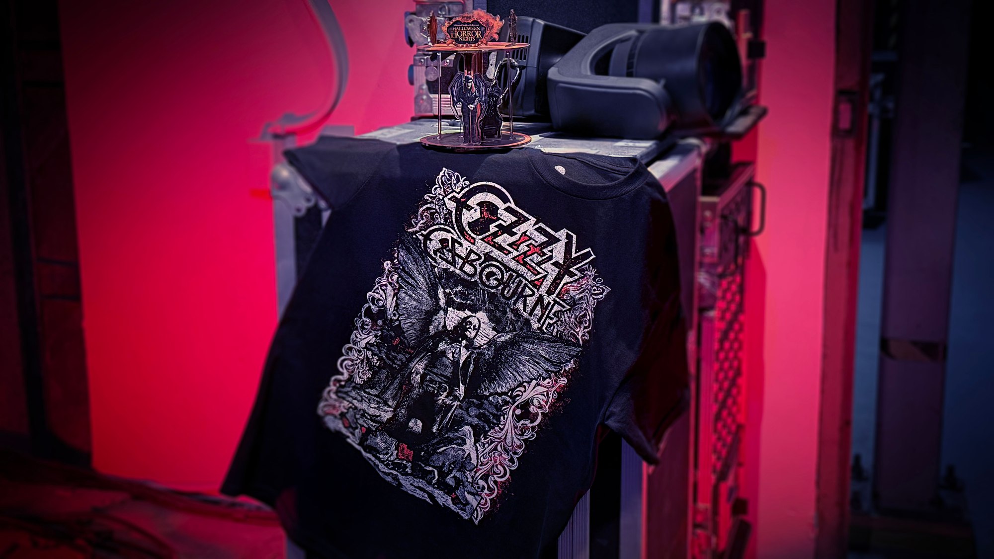 A black Ozzy Osbourne t-shirt draped over a road case in red light, with a small acrylic figure of a winged Ozzy standing and leaning on a cane in front of a throne, displayed on a tiered Halloween Horror Nights stand above it
