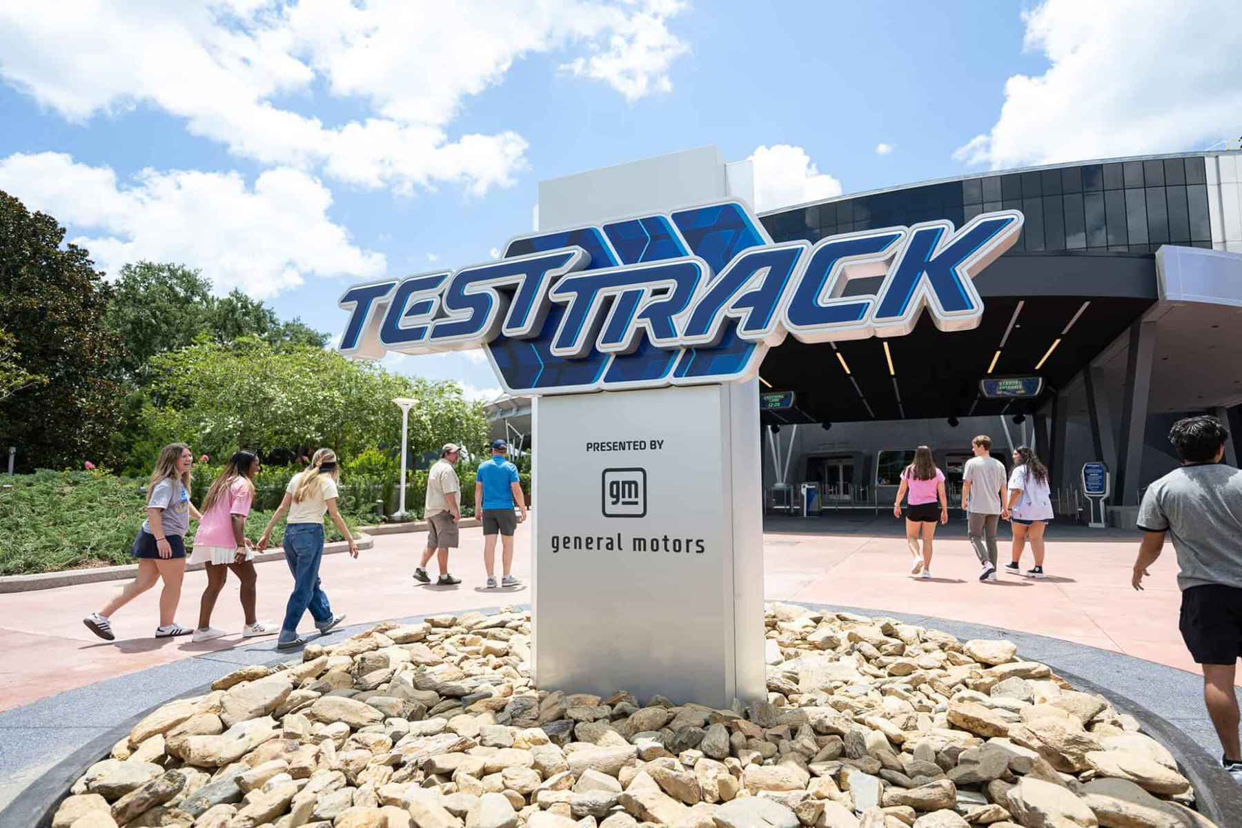 The Test Track marquee outside the EPCOT attraction on a sunny day, with the ride logo above a sign reading Presented By General Motors and guests walking past