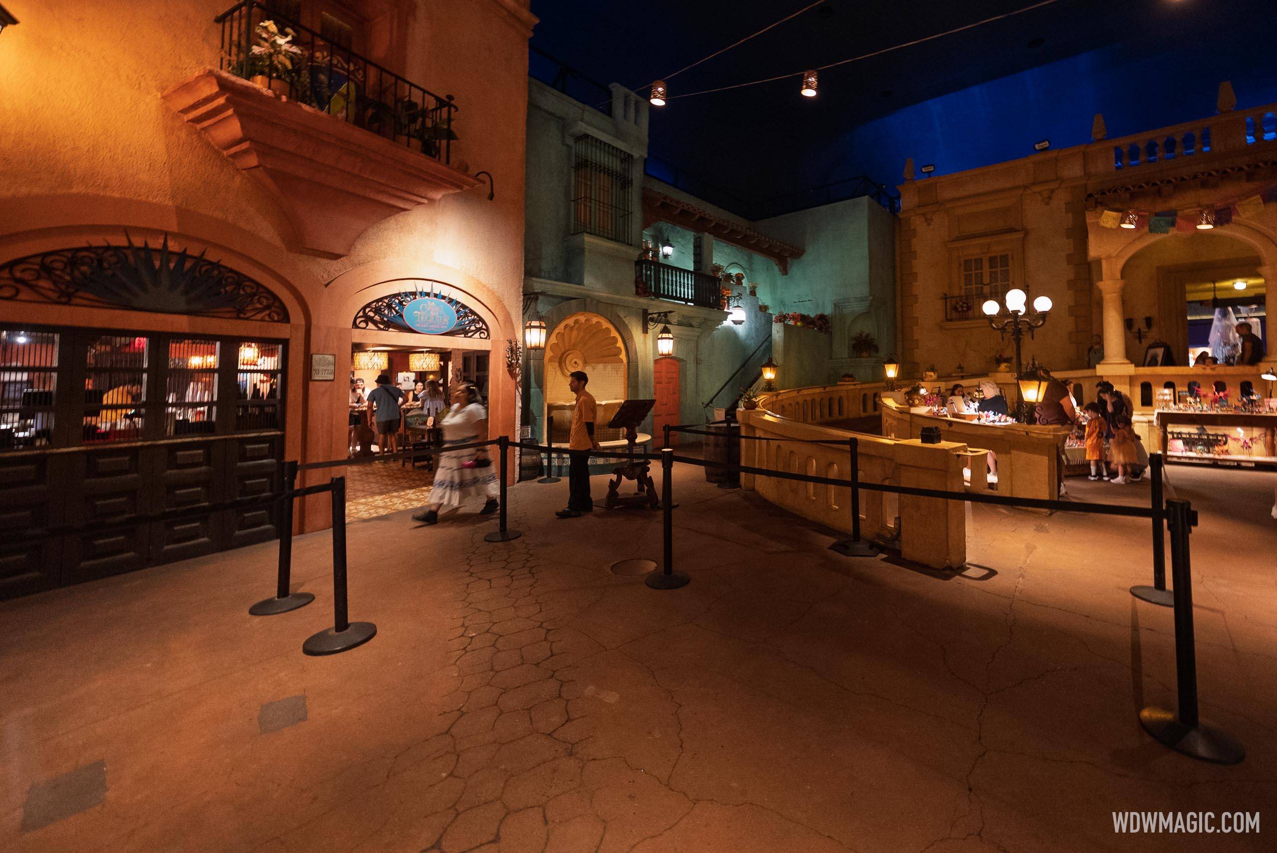 The plaza floor inside the Mexico pavilion looking toward the La Cava del Tequila entry arch with its round blue agave sign, a cast member in a yellow shirt standing at a wooden podium, black retractable belt stanchions zigzagging across the empty foreground pavement, and the carving tables visible on the raised ledge at right