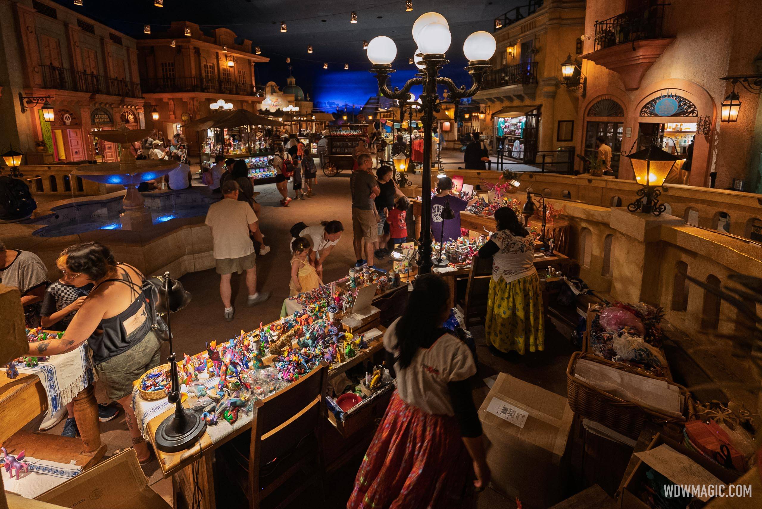 An elevated wide view down a row of long wooden tables of painted carvings on a raised ledge inside the Mexico pavilion marketplace, vendors in embroidered blouses and long patterned skirts working the tables while guests browse, the lit fountain and the La Tienda Encantada storefront visible in the plaza below, and cardboard shipping boxes on the floor beside the tables