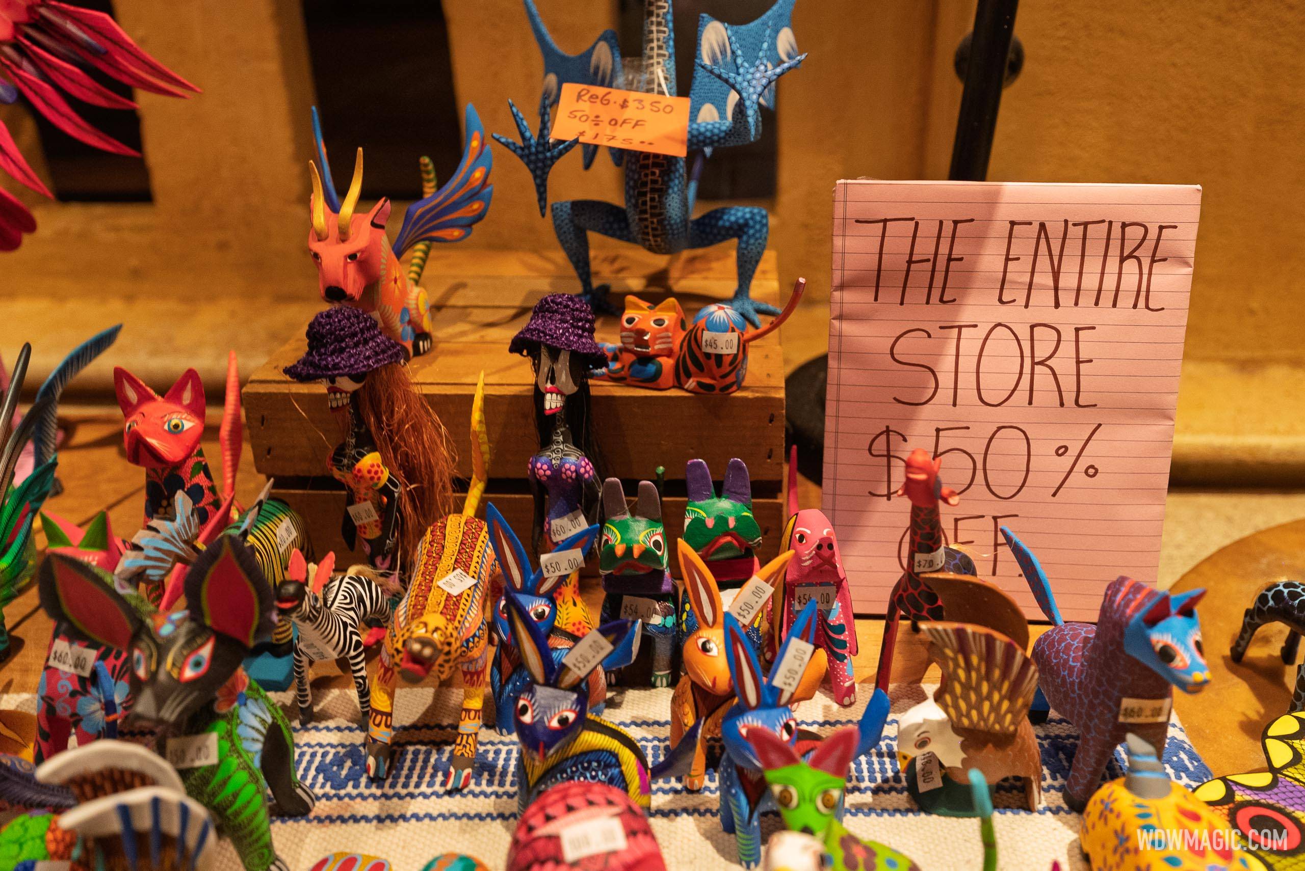 A crowded tabletop of small brightly painted copal wood animals including a red winged wolf, a tall red and black giraffe, a zebra, rabbits and two skeleton figures in purple straw hats, with white price tags reading 45 and 50 and 54 and 60 and 70 dollars, a blue spotted lizard mounted behind them carrying an orange tag reading REG. $350, 50% OFF, $175.00, and a sheet of pink lined notepad paper propped upright, hand lettered THE ENTIRE STORE $50% OFF