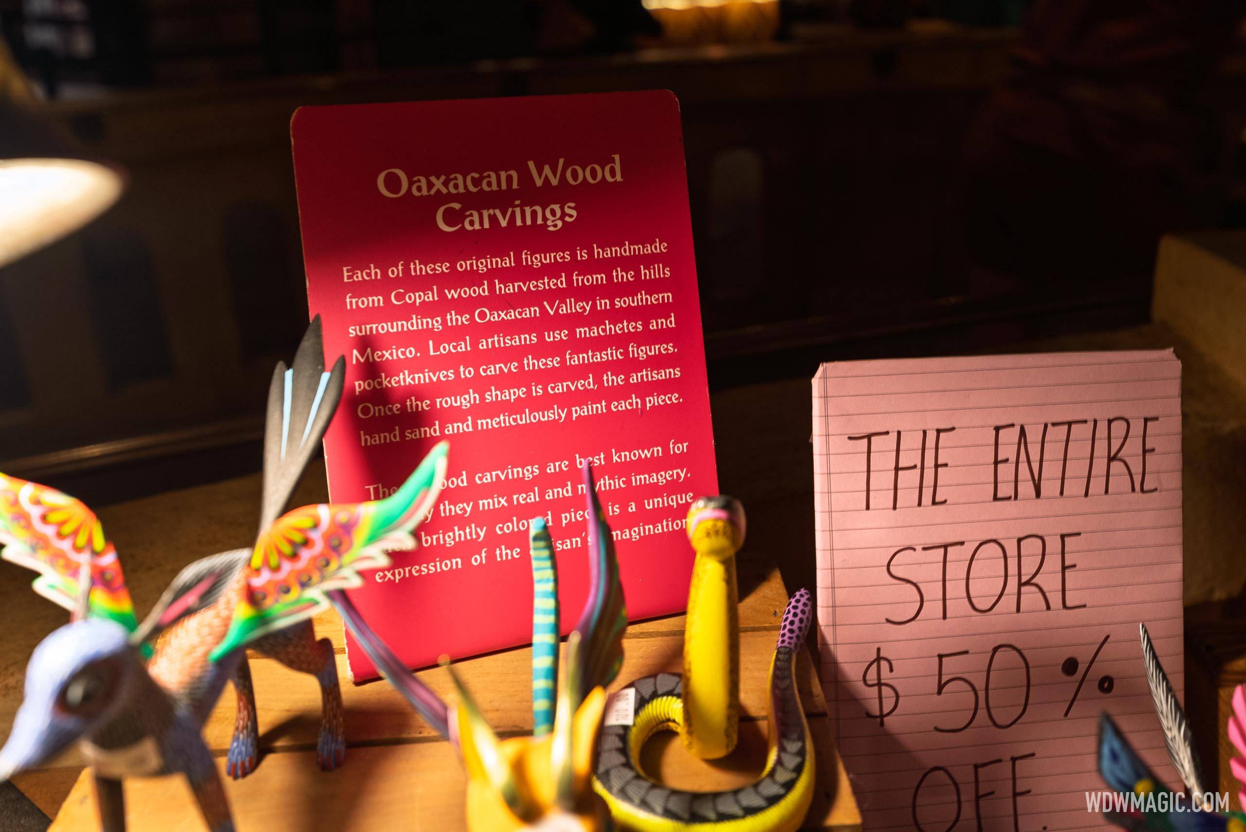 A red interpretive card headed Oaxacan Wood Carvings in gold lettering, standing on a wooden crate with its first paragraph legible, a long snouted four legged animal with large rainbow eye spotted wings and a coiled yellow and grey banded snake in the foreground, and the handwritten pink 50 percent off notepad sign standing to its right