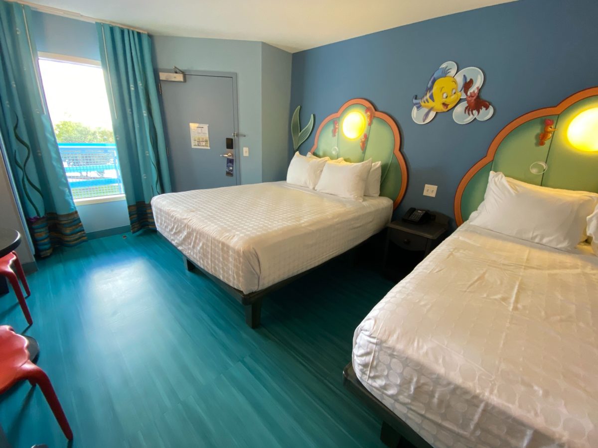 A Little Mermaid standard room with two beds, green and orange scallop shell headboards lit by round pearl lights, a Flounder and Sebastian wall cutout, teal curtains and glossy teal wood look flooring