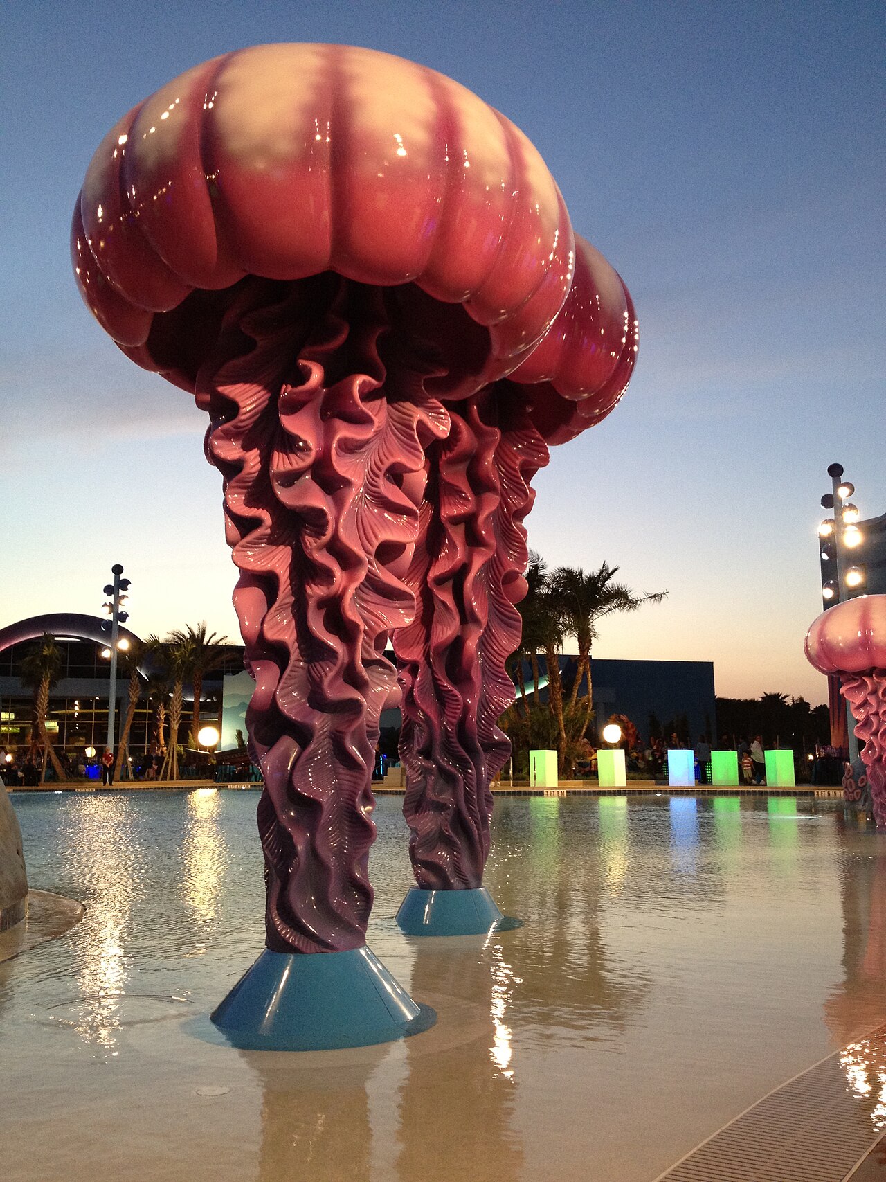 Large pink and purple jellyfish sculptures with ruffled tentacles standing in the shallow end of a pool at dusk, with palm trees, glowing green and blue light boxes and resort buildings behind