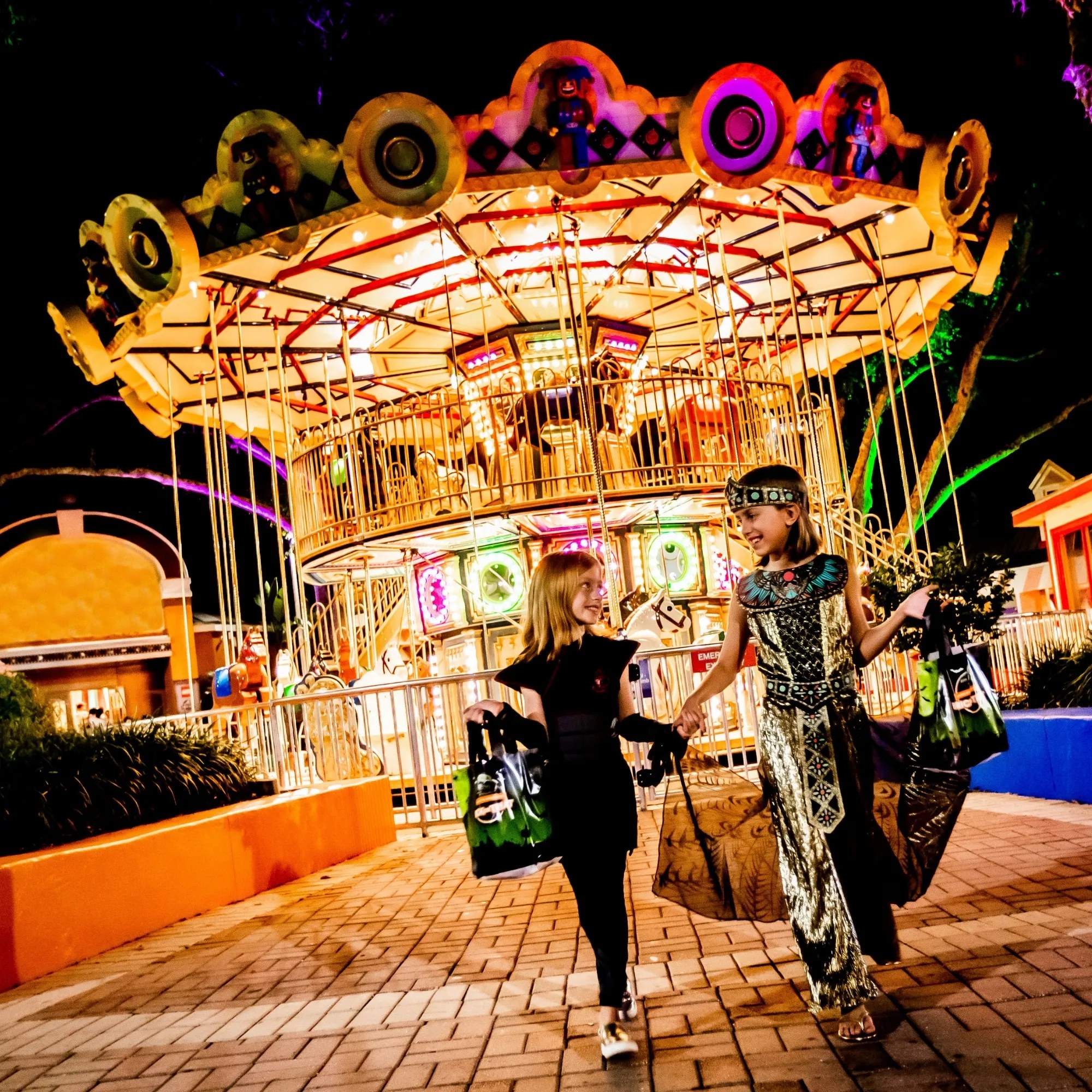 A double decker carousel lit gold at night with purple and green light washing the trees behind it, and two girls in Halloween costumes walking toward the camera holding hands and carrying green and black trick or treat bags