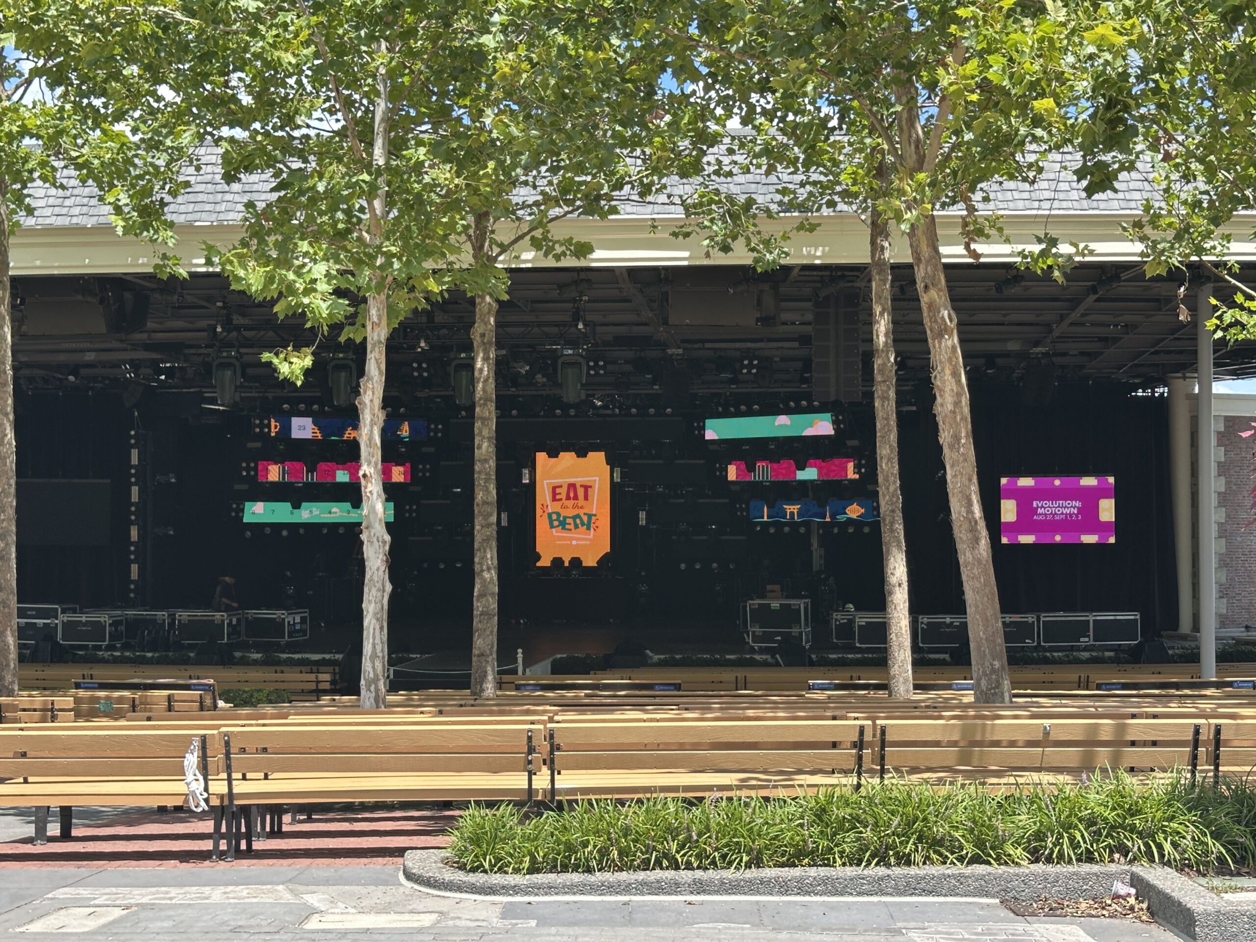 The open-air America Gardens Theatre stage at EPCOT seen through the trees, with an orange Eat to the Beat ticket graphic on the black LED wall, road cases along the stage floor and empty wooden bench seating in front