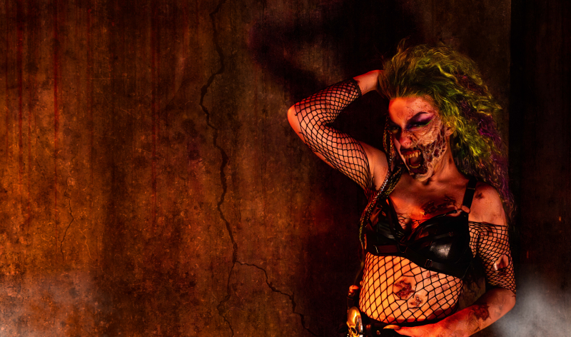 A Howl-O-Scream scare actor with long green and yellow hair, torn prosthetic makeup across the mouth and jaw, fishnet sleeves and a leather harness, posed against a cracked rust colored concrete wall
