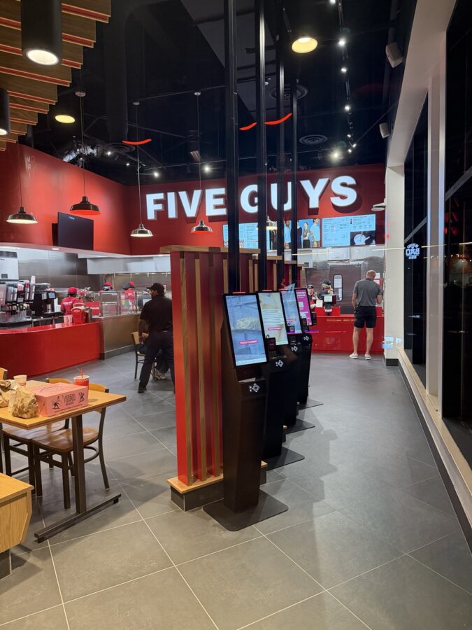 The finished interior of a Five Guys restaurant, with a lit white FIVE GUYS wall sign on a red back wall, digital menu boards, four black self order kiosks in a row on grey tile, a wooden slat divider, wood tables and chairs, several employees in red behind a red service counter, a man in a black shirt and black cap beside a chair in the dining area and another guest standing at the counter on the right