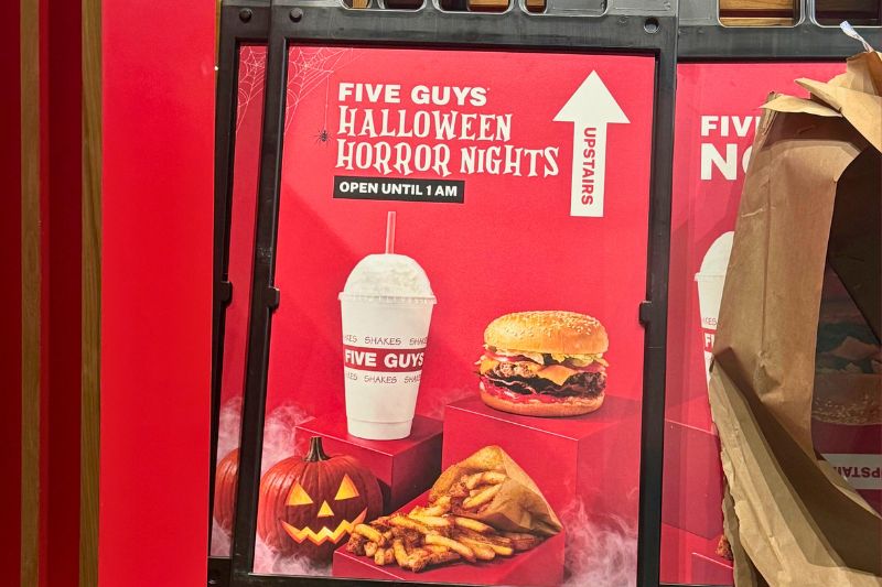 A red poster behind glass in a black metal frame reading FIVE GUYS, then HALLOWEEN HORROR NIGHTS, then OPEN UNTIL 1 AM in a black bar, with a white up arrow and vertical UPSTAIRS text at the right, a cobweb and a small dangling spider at the left, and below them a milkshake cup and a cheeseburger on red risers beside a carved jack o lantern and a paper wrap of fries, all in fog