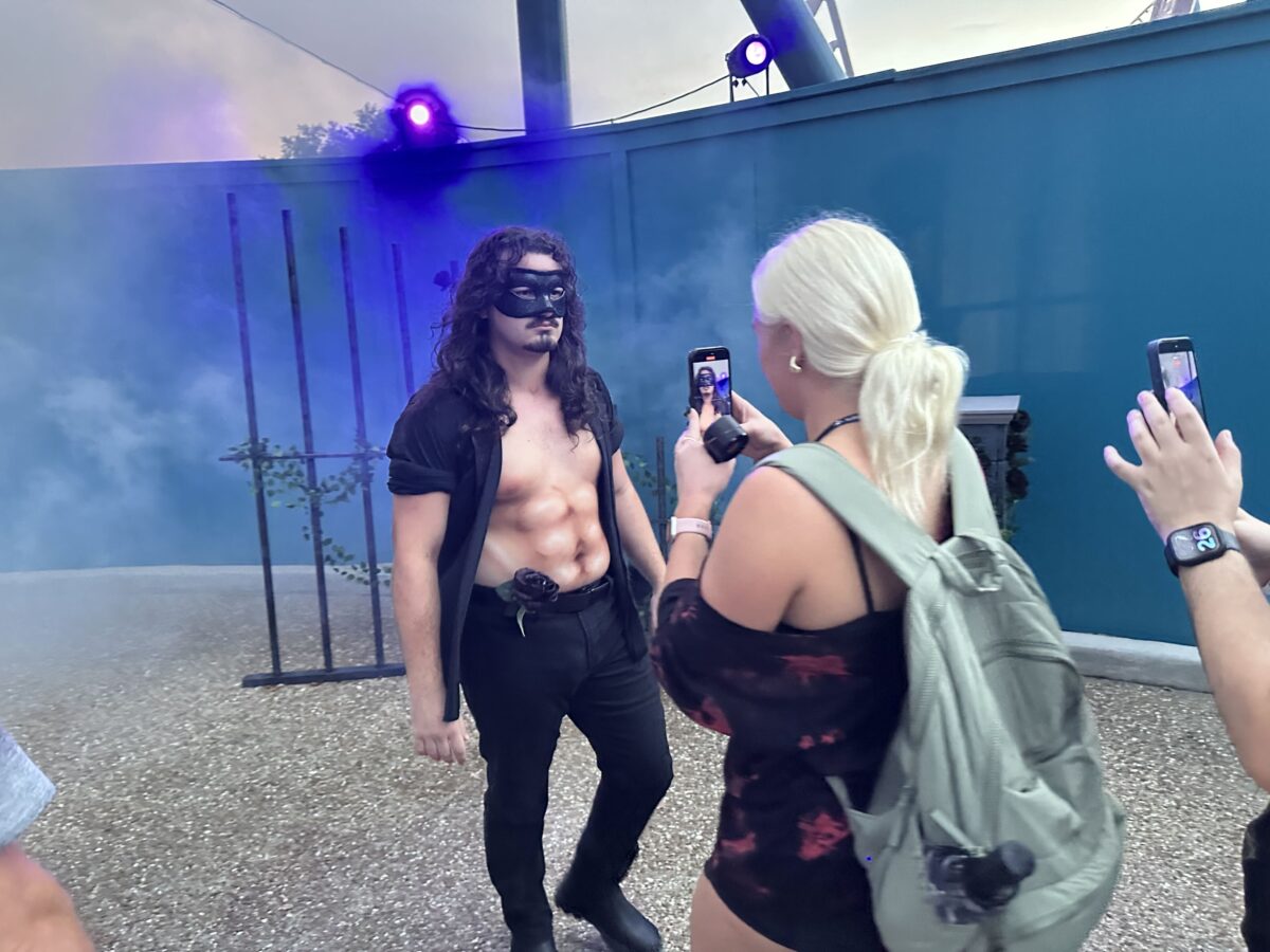 A shirtless scare actor with long dark hair and a black domino mask, wearing an open black shirt and black jeans with a black rose tucked into his belt, standing in purple lit fog beside a black wrought iron fence, while a blonde woman with a green backpack films him on a phone in the right foreground