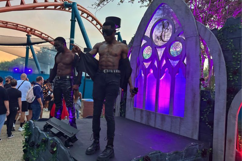 Two masked scare actors in black jeans and boots standing on a low stage, both bare chested in open black jackets, beside a grey stone gothic arch with a stained glass window lit magenta and purple, two rose medallions and a JOMC monogram in the tracery, ivy trailing over the stonework, with an orange roller coaster track on teal supports and a crowd of guests to their left at dusk