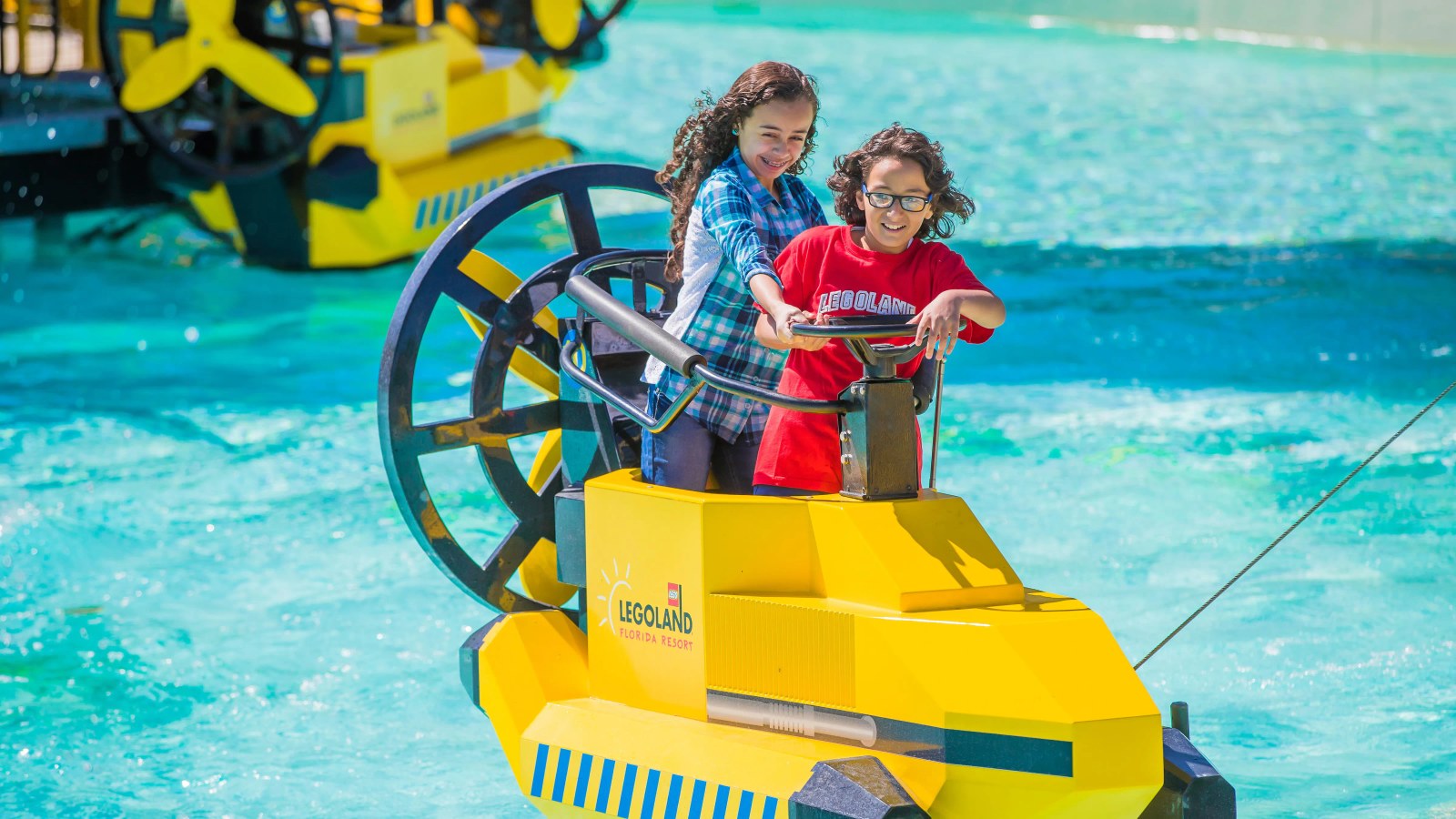 Two children standing together on a yellow LEGO styled wave racer craft on bright turquoise water, one in a red LEGOLAND shirt steering, the craft's hull carrying the LEGOLAND Florida Resort sun logo, with a large black and yellow spoked wheel behind them