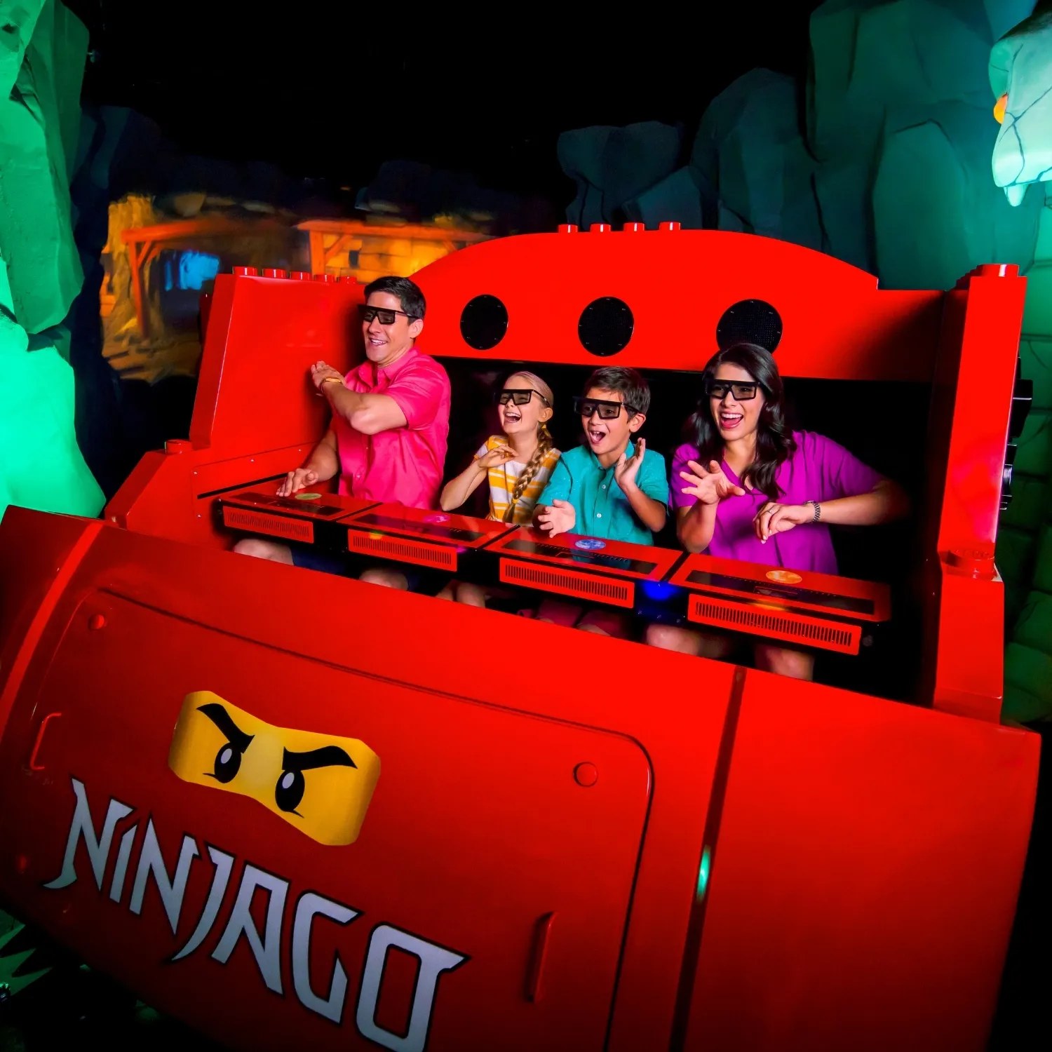 A four person family in black 3D glasses riding a bright red NINJAGO ride car through a dark show scene, the car front carrying yellow ninja eyes and the NINJAGO wordmark, with glowing green rock formations and an orange lit structure behind, all four riders with hands raised