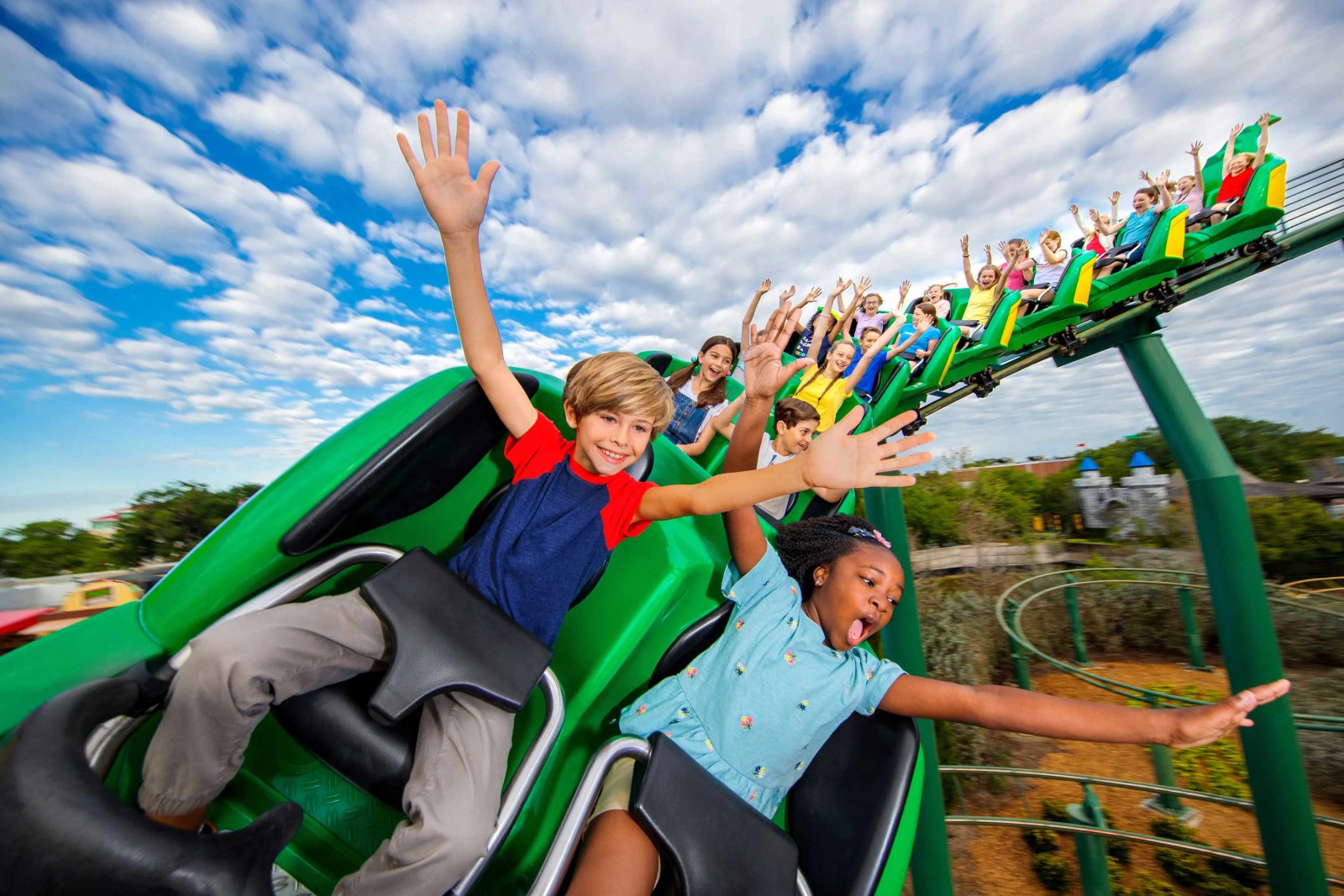 A green coaster train full of children on a section of outdoor track under a blue sky with mottled cloud, most riders with arms raised and the boy in front with one hand high, with a grey castle facade and landscaped beds visible at right