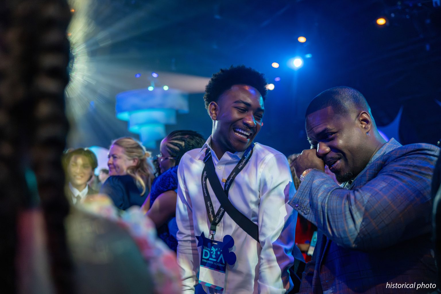 A smiling teenager in a white shirt wearing a Disney Dreamers Academy lanyard and badge stands beside a man in a plaid blazer who wipes his eye, at a dimly lit event with blue stage lights