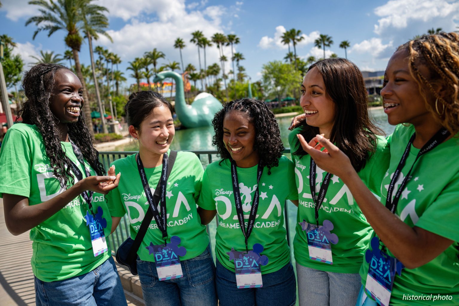 Five smiling teenagers in matching green Disney Dreamers Academy T-shirts and lanyards stand together at a railing, with palm trees and a large green dinosaur figure in a lagoon behind them