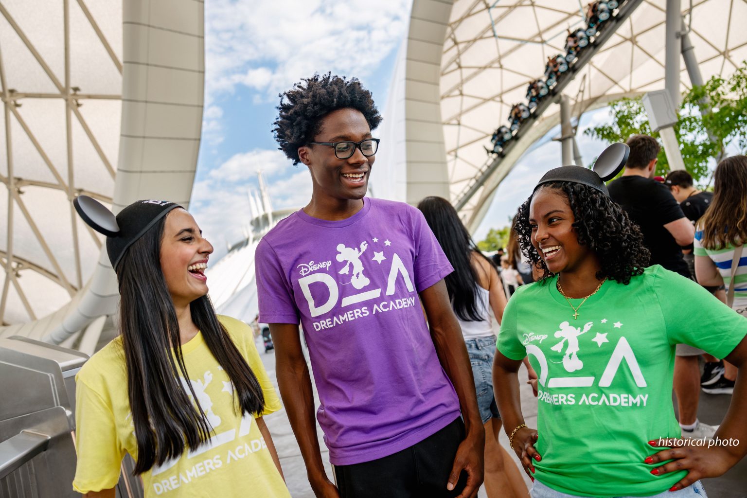 Three smiling teenagers in yellow, purple and green Disney Dreamers Academy T-shirts, two wearing Mickey ears, stand under a white curved canopy with Space Mountain visible in the background