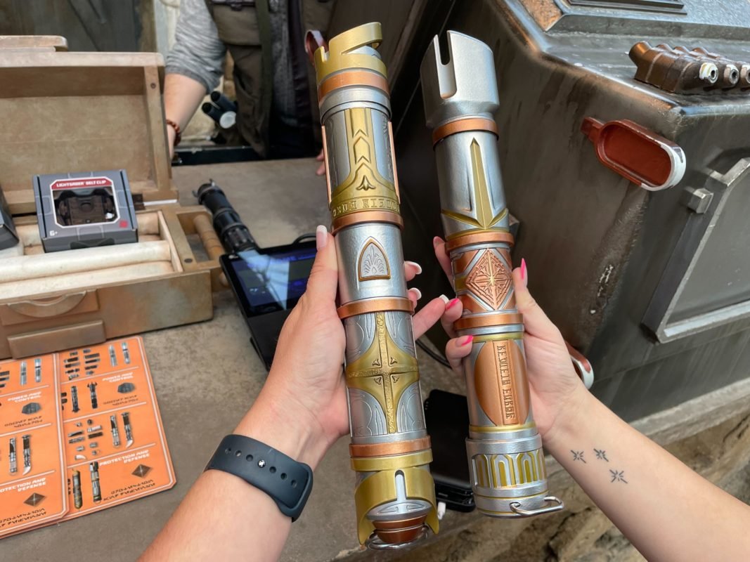 Two hands hold up two silver, gold and copper lightsaber hilts beside a Savi's Workshop workbench with an orange parts card on the counter