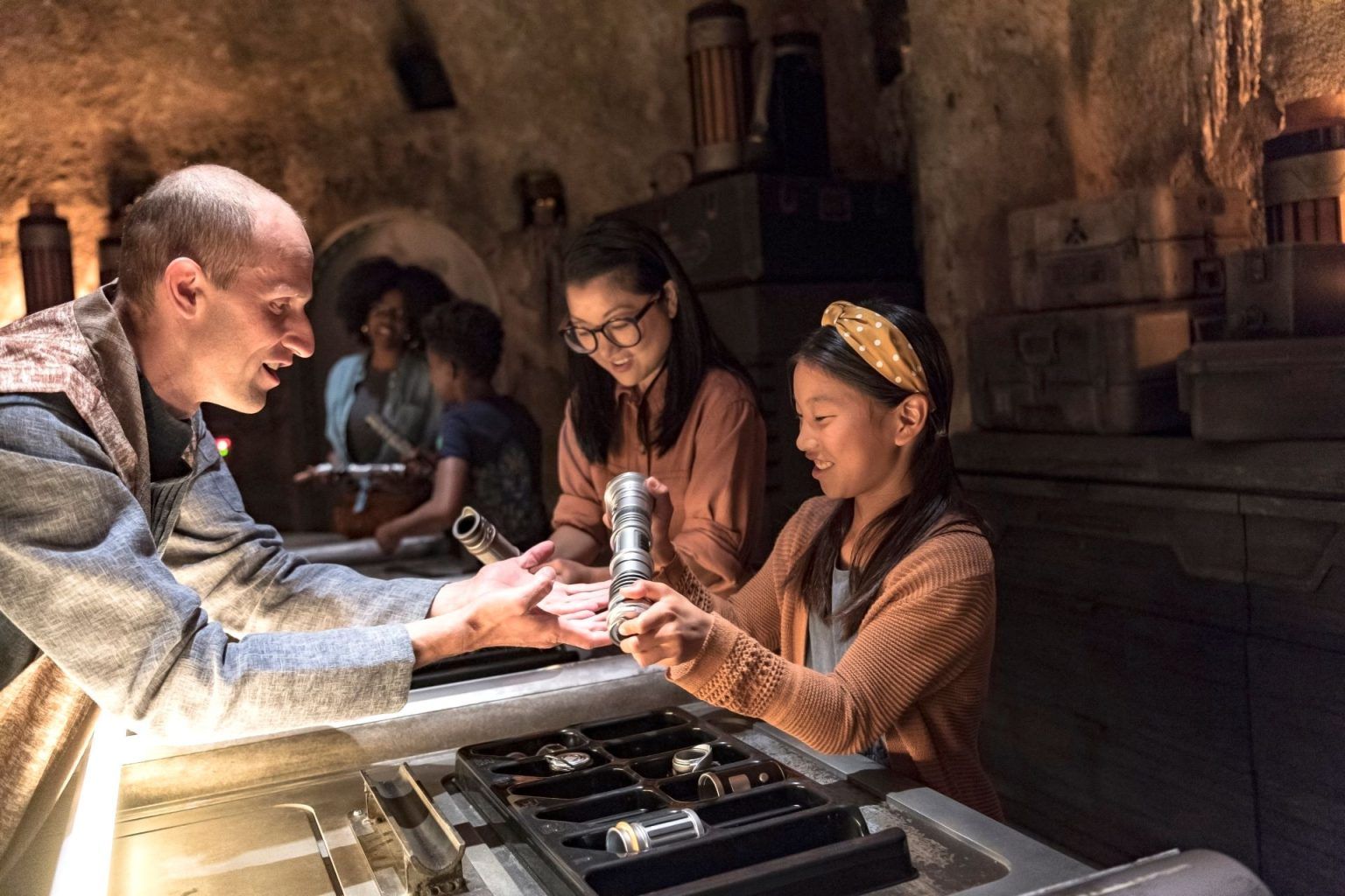 A Savi's Workshop Gatherer holds out his hands as a girl assembles a silver lightsaber hilt over a tray of parts