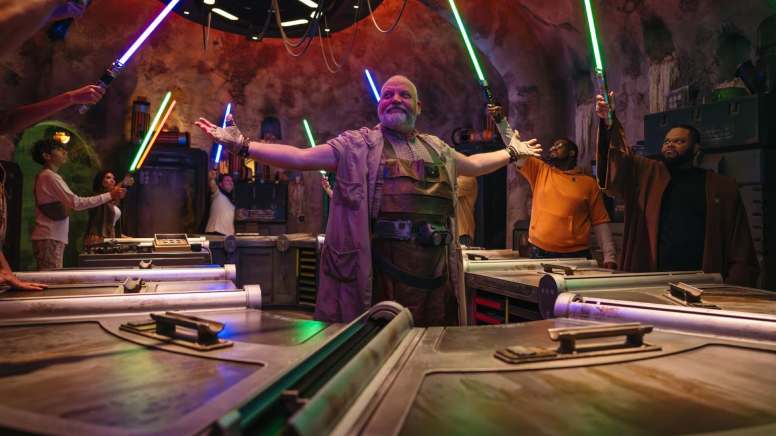 Guests raise lit blue, green, violet and orange lightsabers around the workbenches inside Savi's Workshop as a Gatherer spreads his arms in the center