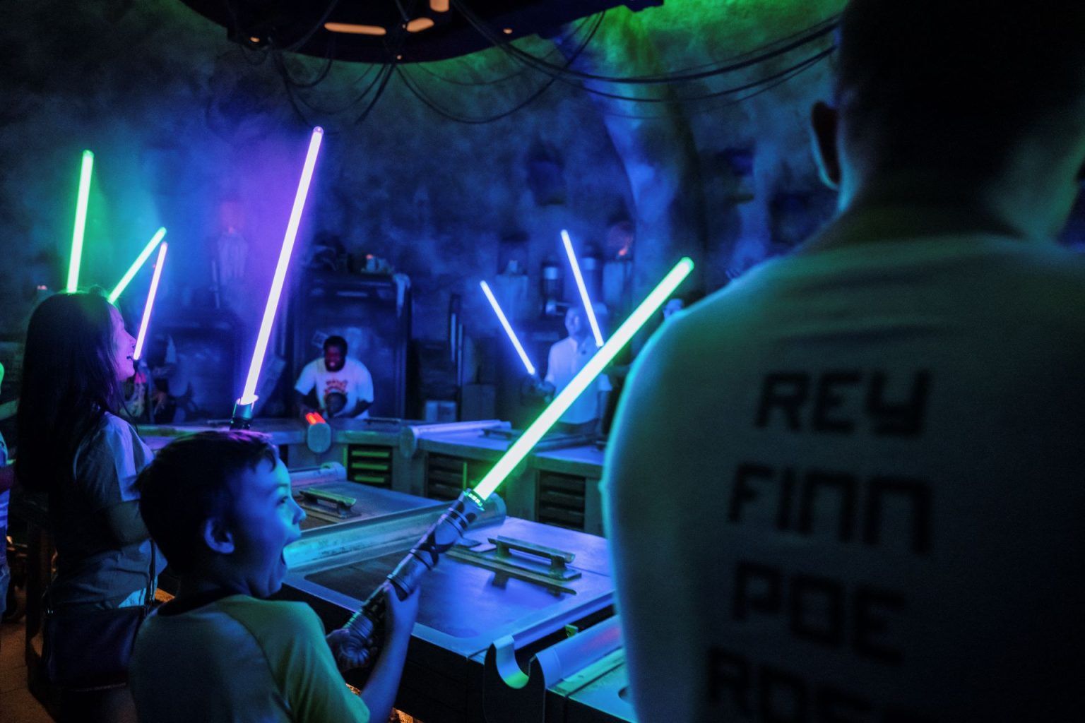 A boy holds a lit green lightsaber at a Savi's Workshop workbench while other builders raise green, blue and violet blades in the background