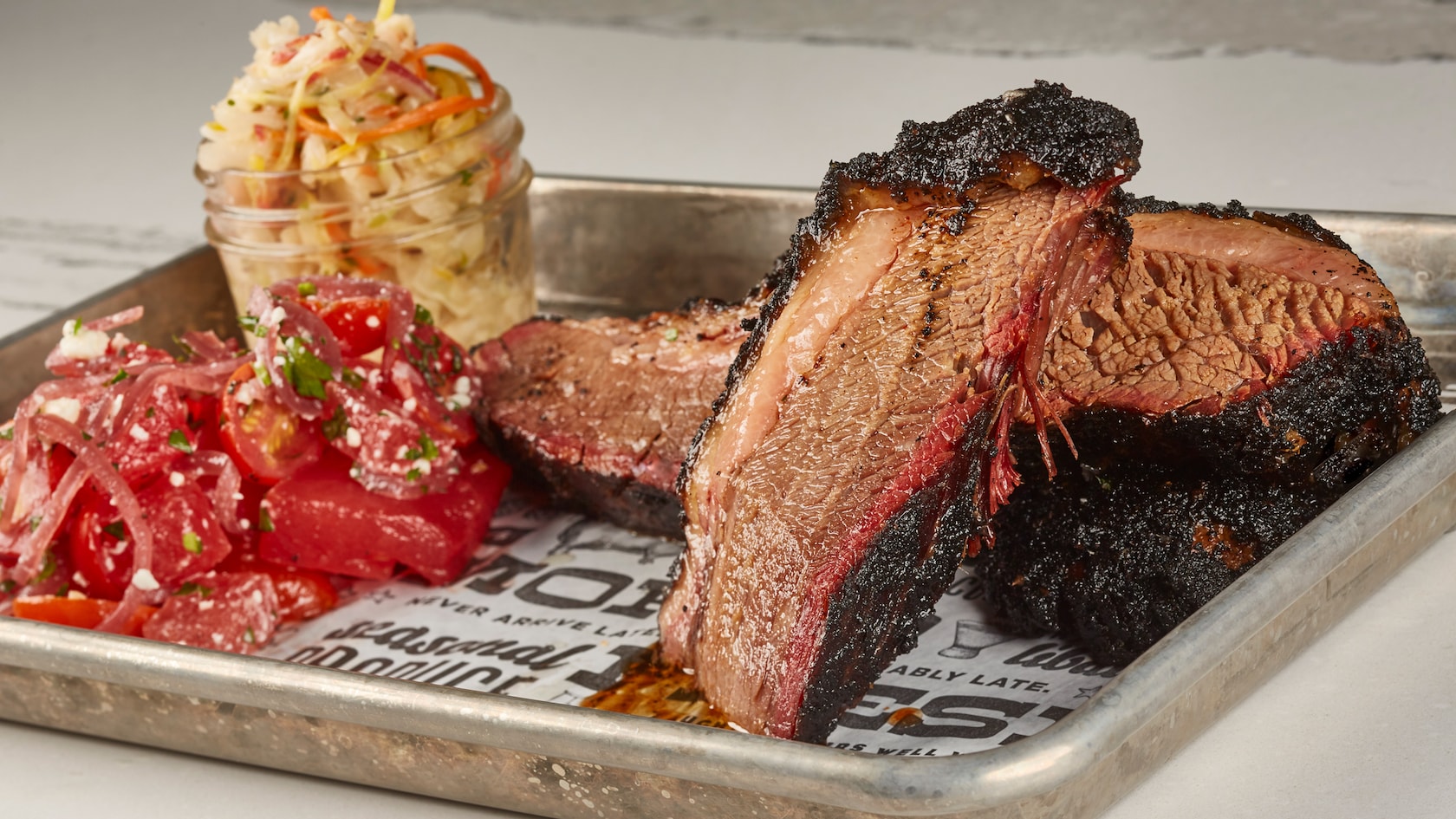 Sliced smoked brisket with a dark peppery bark on a metal tray, beside a watermelon and tomato salad and a jar of slaw