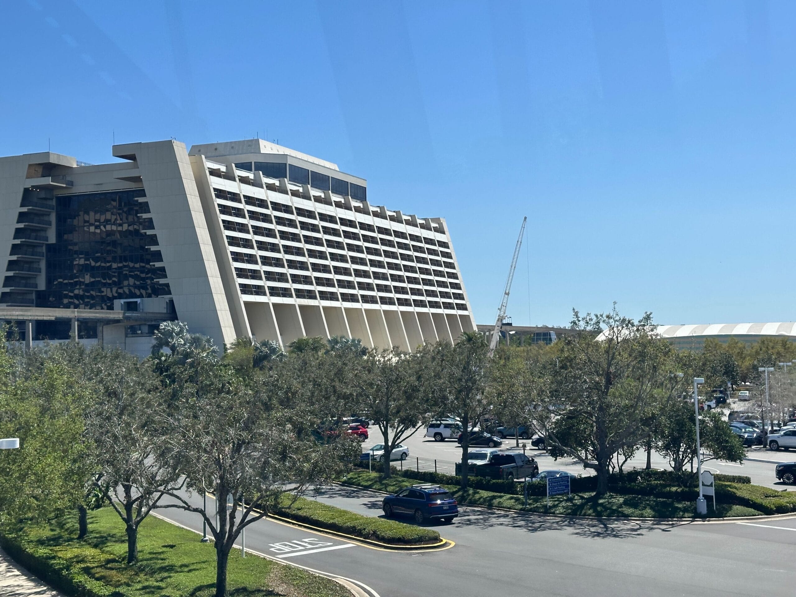 The A-frame tower of Disney's Contemporary Resort under a clear blue sky, with a construction crane rising beyond its right end, and trees and a parking lot in the foreground