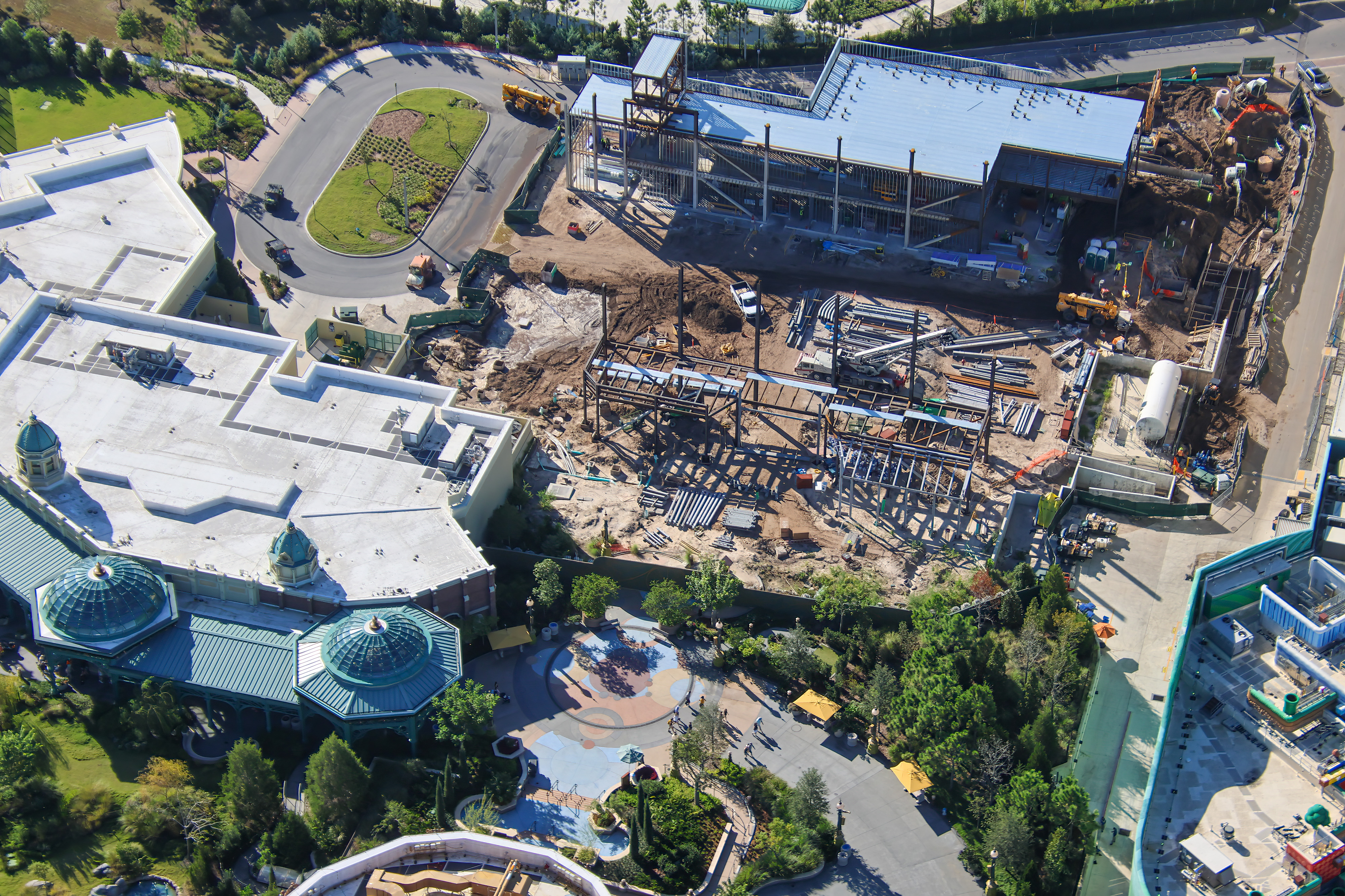 Aerial view looking down on the events building construction site, with a roofed steel structure, open steel framing and stacked materials, beside a large building with green glass domes, above a Celestial Park plaza, with the edge of SUPER NINTENDO WORLD at right